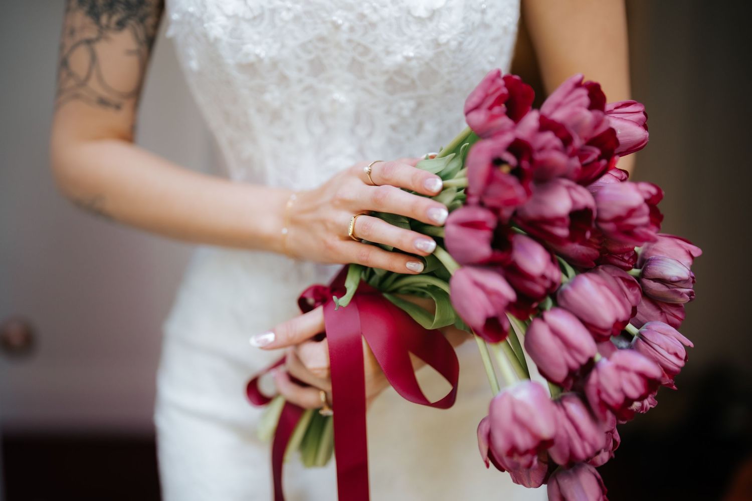 Bride in white lace dress holding a bouquet of deep pink tulips tied with a satin ribbon, wearing a gold ring.