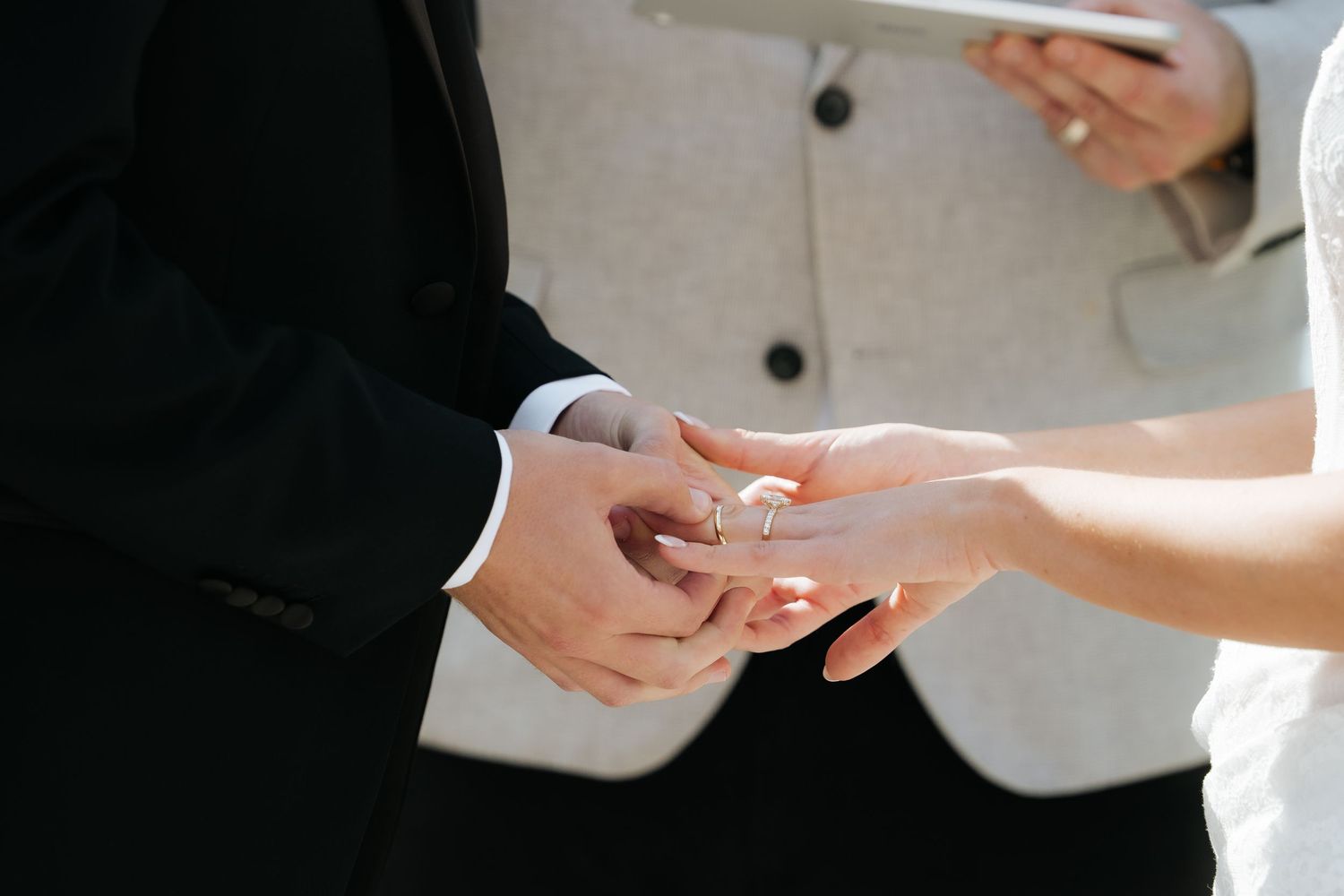 Groom placing a wedding ring on bride's finger during outdoor ceremony, officiant in gray suit visible behind.