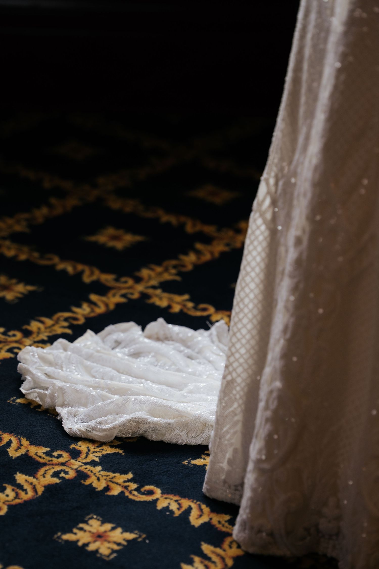 Close-up of intricate lace wedding dress train pooling on ornate dark patterned carpet.
