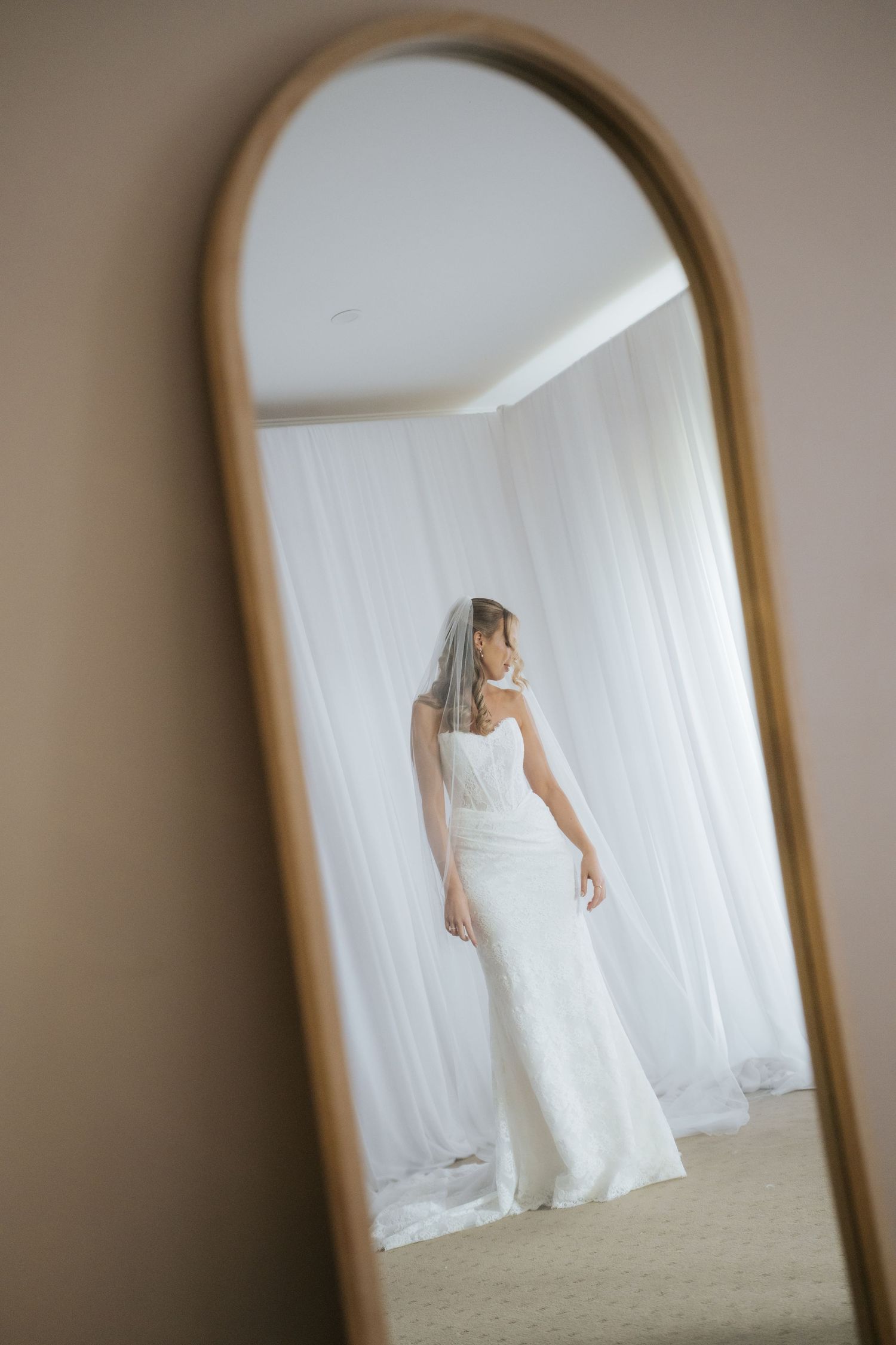 Bride in white wedding gown and veil viewed from behind in arched gold mirror reflection against sheer white curtains.