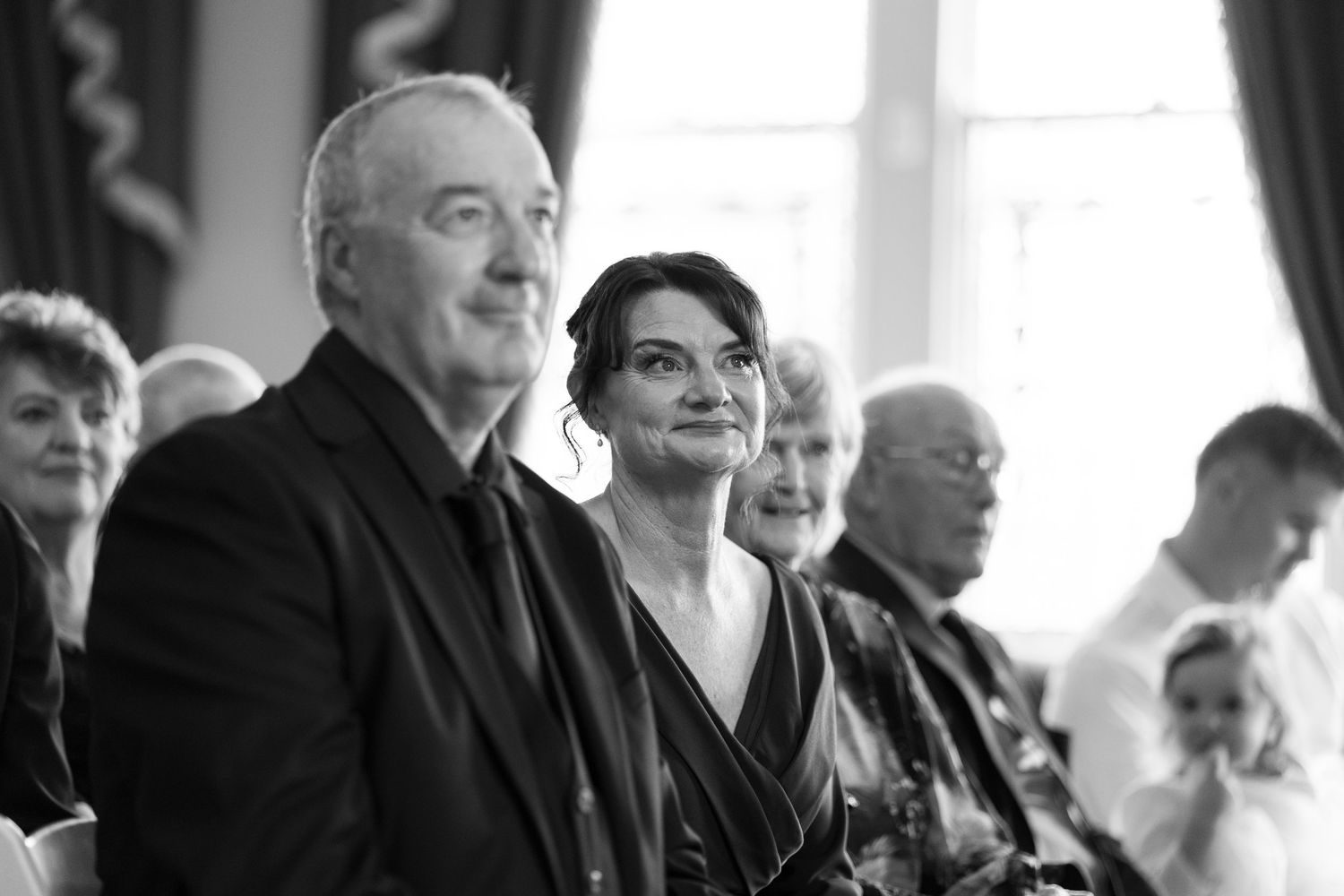 Black and white photo of guests smiling during a wedding ceremony seated in elegant venue.