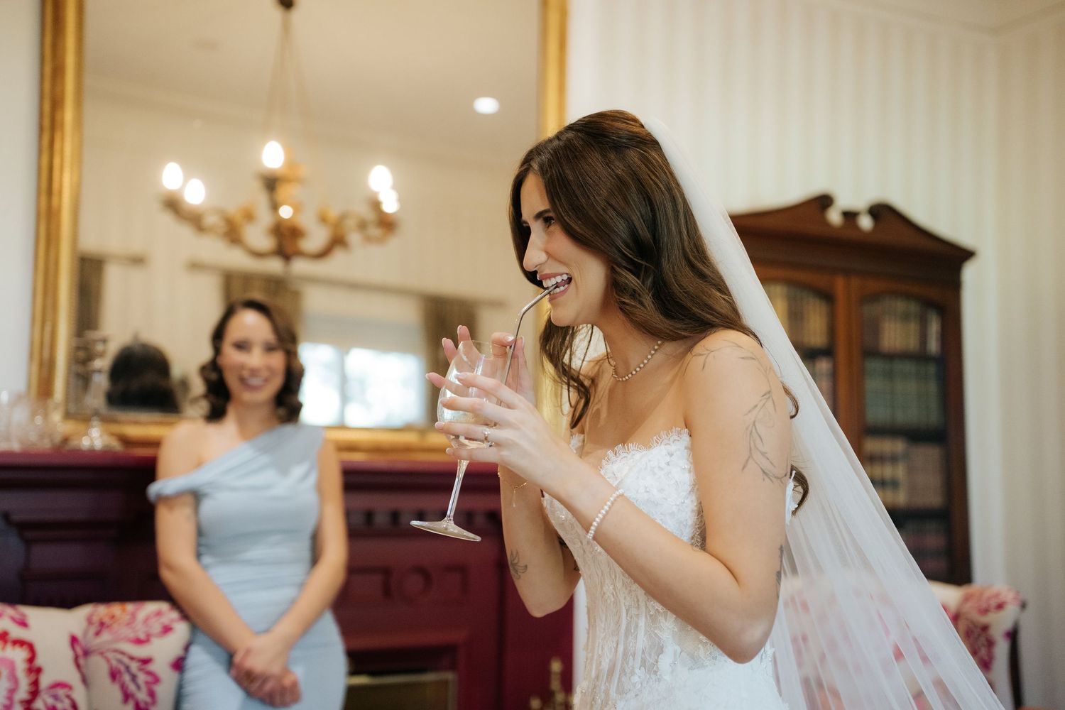 Bride in white gown holding champagne glass laughing during wedding preparations with guest in blue dress.