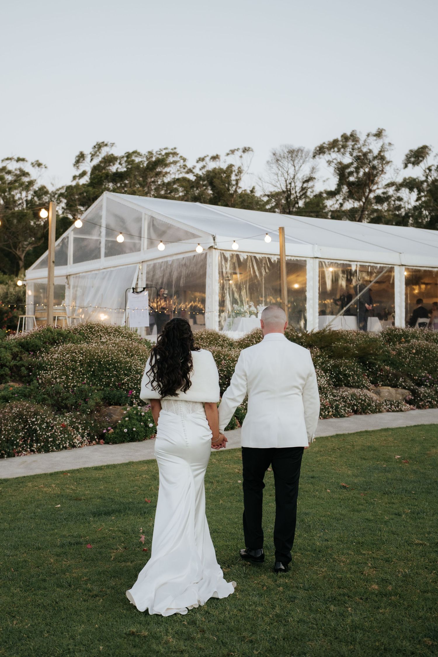 Bride and groom holding hands walking toward illuminated clear wedding reception tent at dusk.