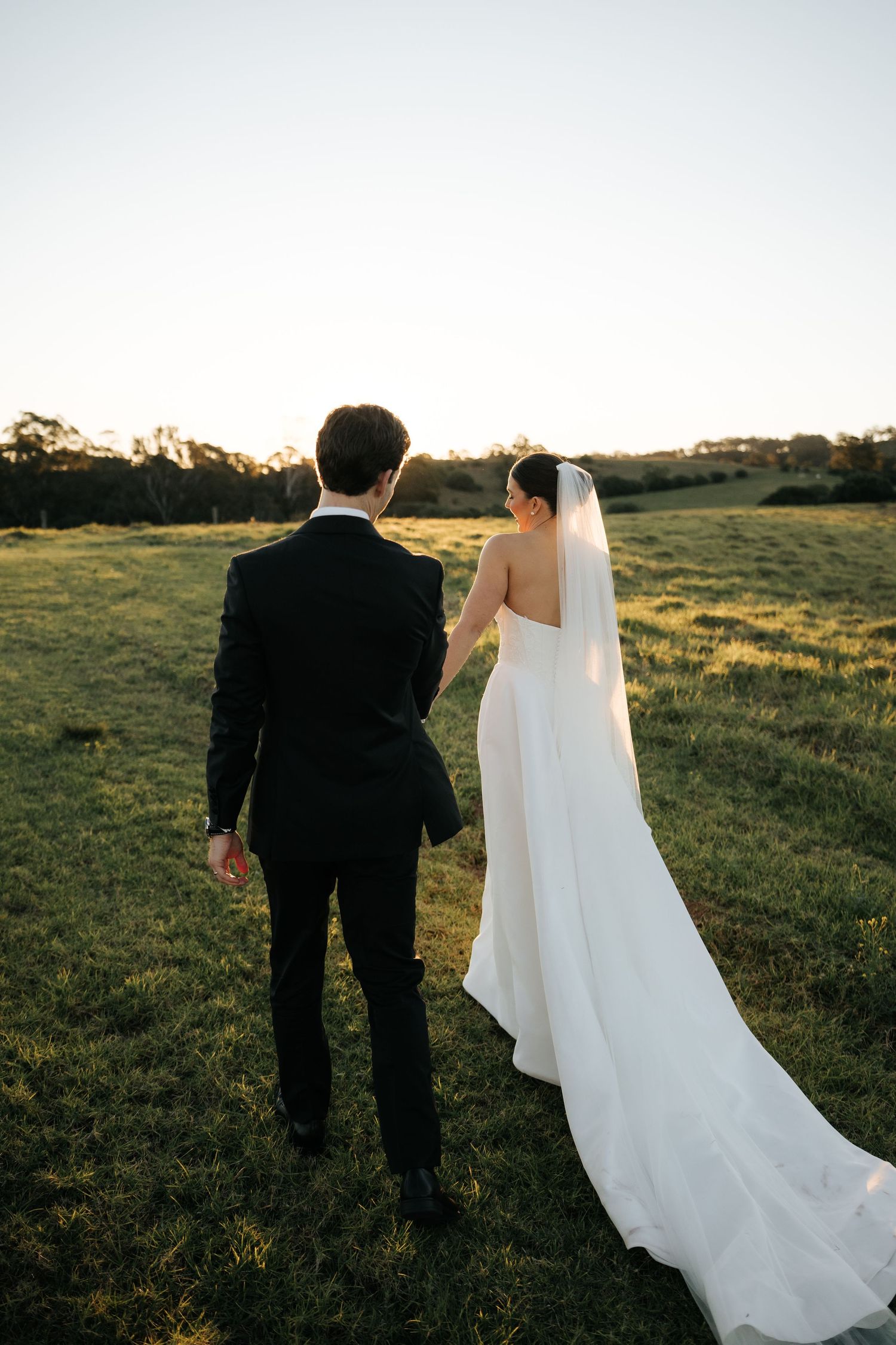 Bride and groom walking hand in hand across green field at golden hour sunset.