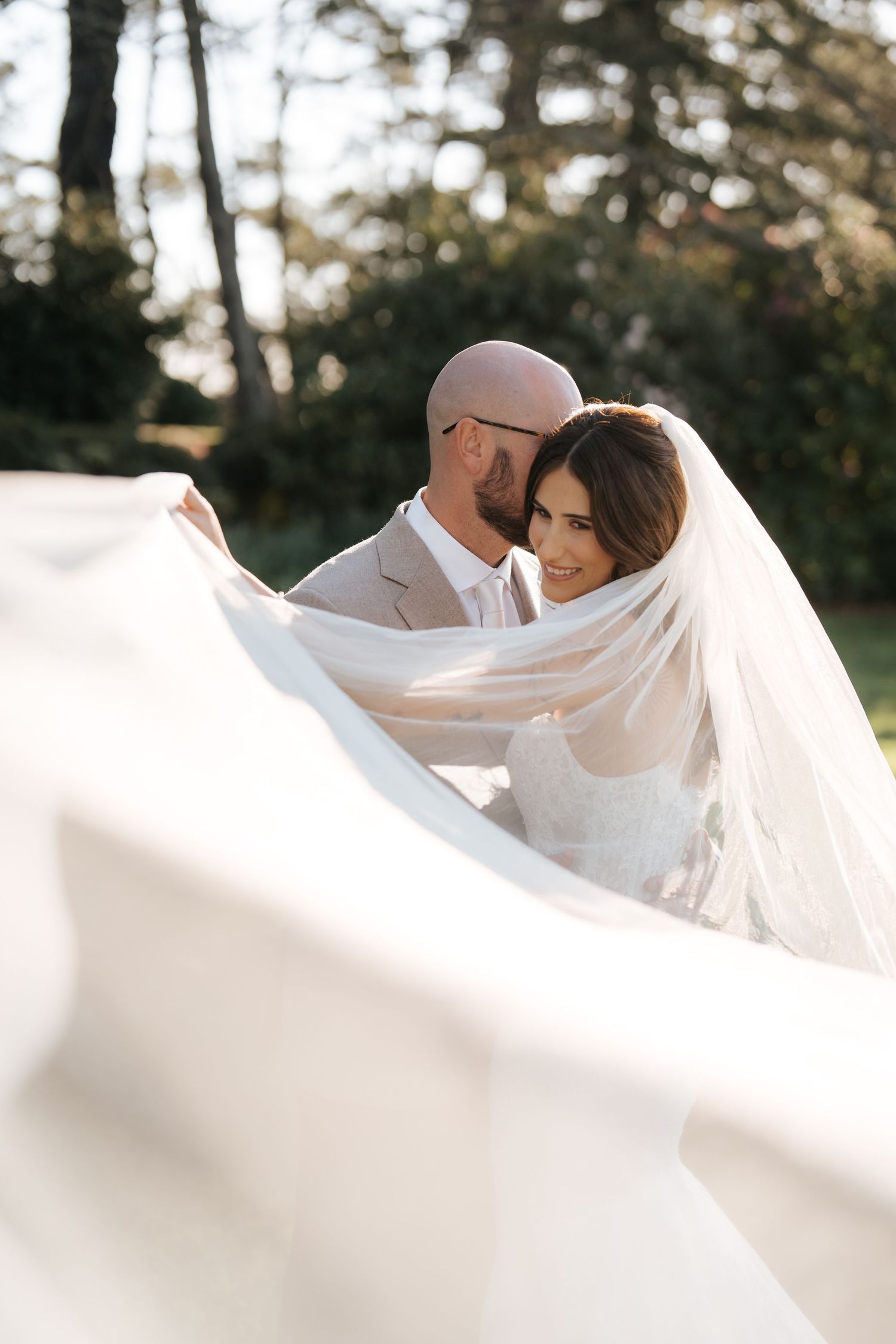 Bride and groom embrace outdoors, her flowing veil billowing in the breeze, surrounded by lush green trees.