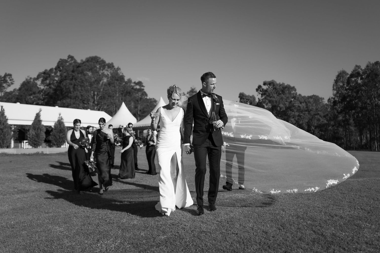 Black and white wedding photo of bride and groom walking on grass with bridal party and marquee in background.