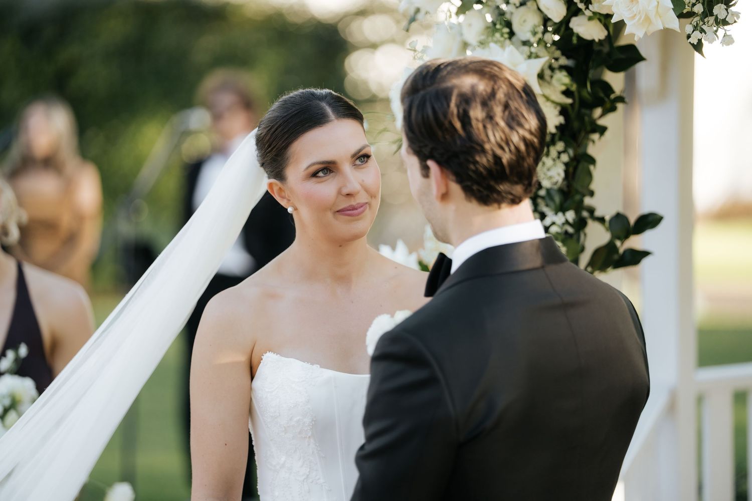 Bride smiling at groom during outdoor wedding ceremony with white floral arch backdrop.