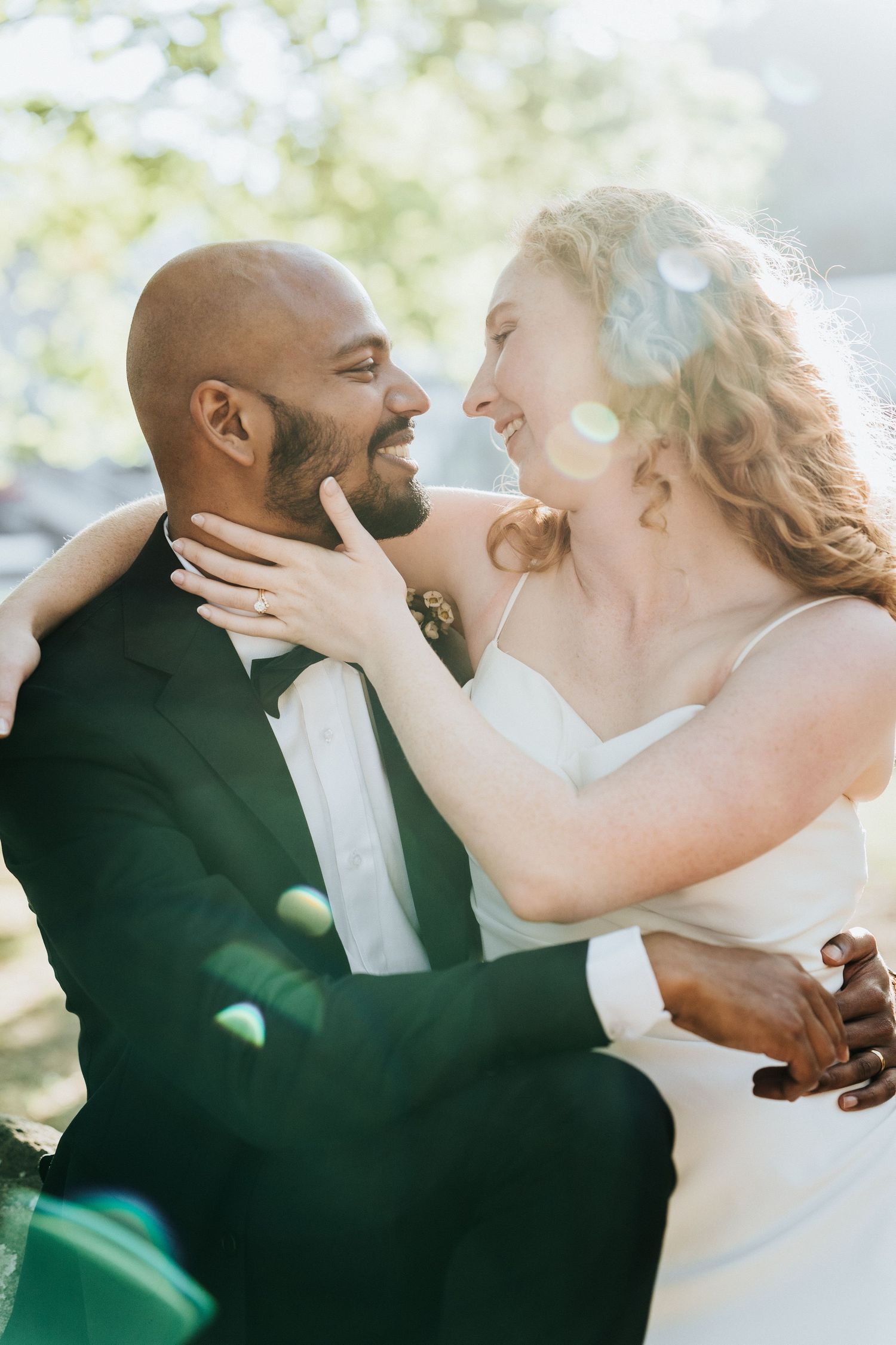 A joyful interracial couple in wedding attire share a romantic moment, smiling at each other in soft sunlight.
