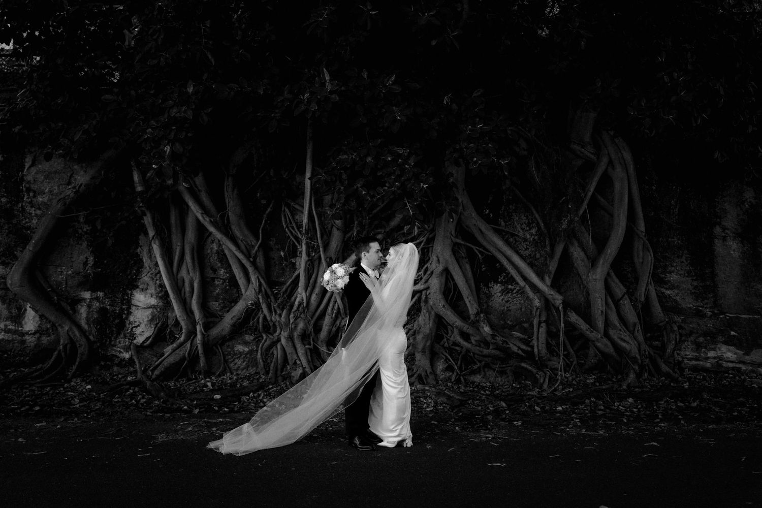Black and white photo of a bride and groom embracing before dramatic twisted tree roots at night.