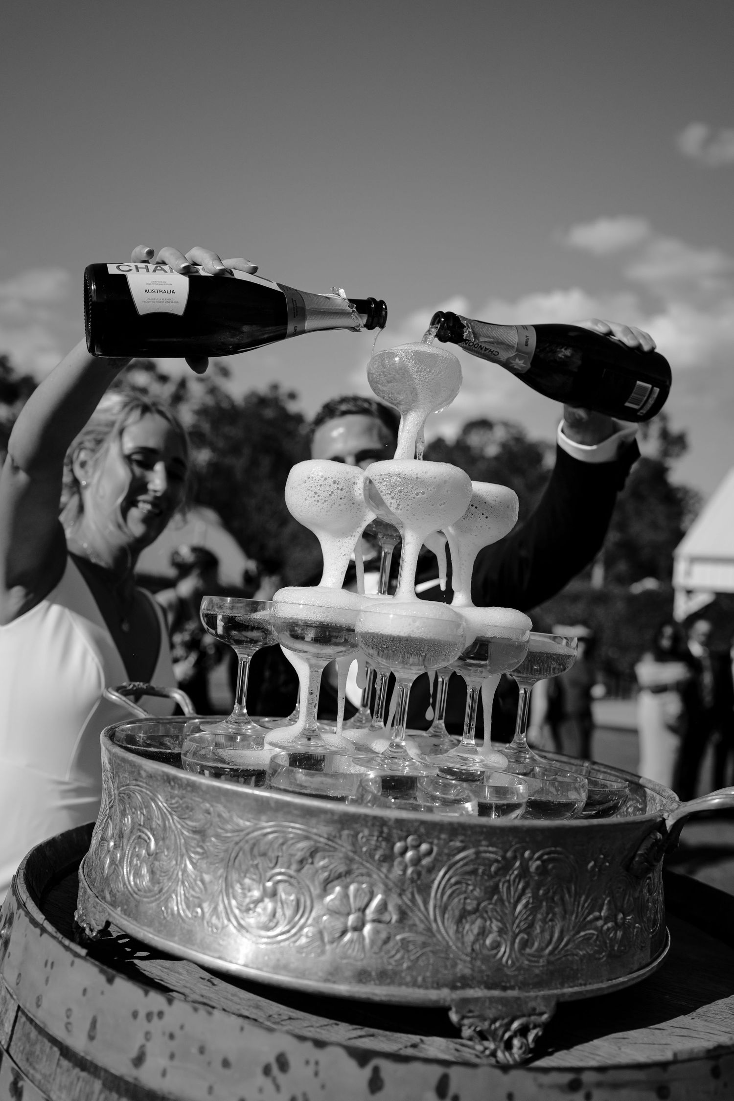Black and white photo of champagne tower being filled with bottles at an outdoor wedding celebration.