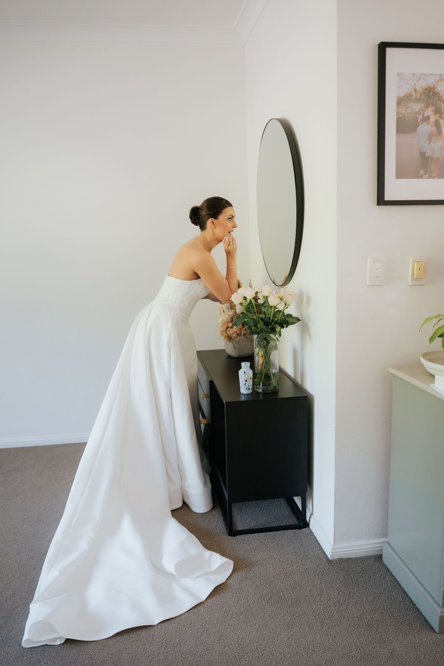 Bride in strapless white gown checking her makeup in a round mirror above a black dresser with fresh flowers.
