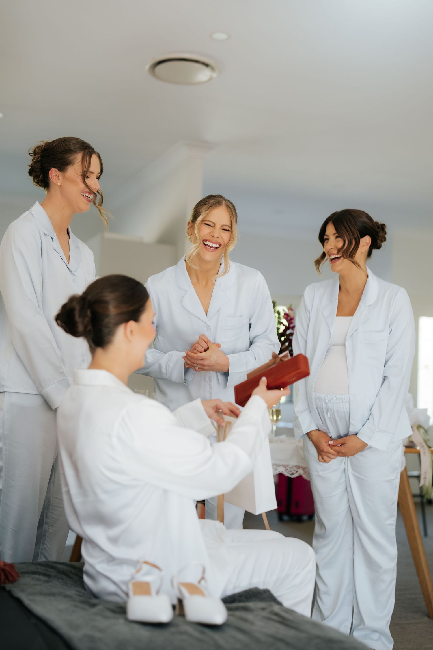 Bridesmaids in matching light blue pajamas laughing and sharing gifts during bridal morning preparations.