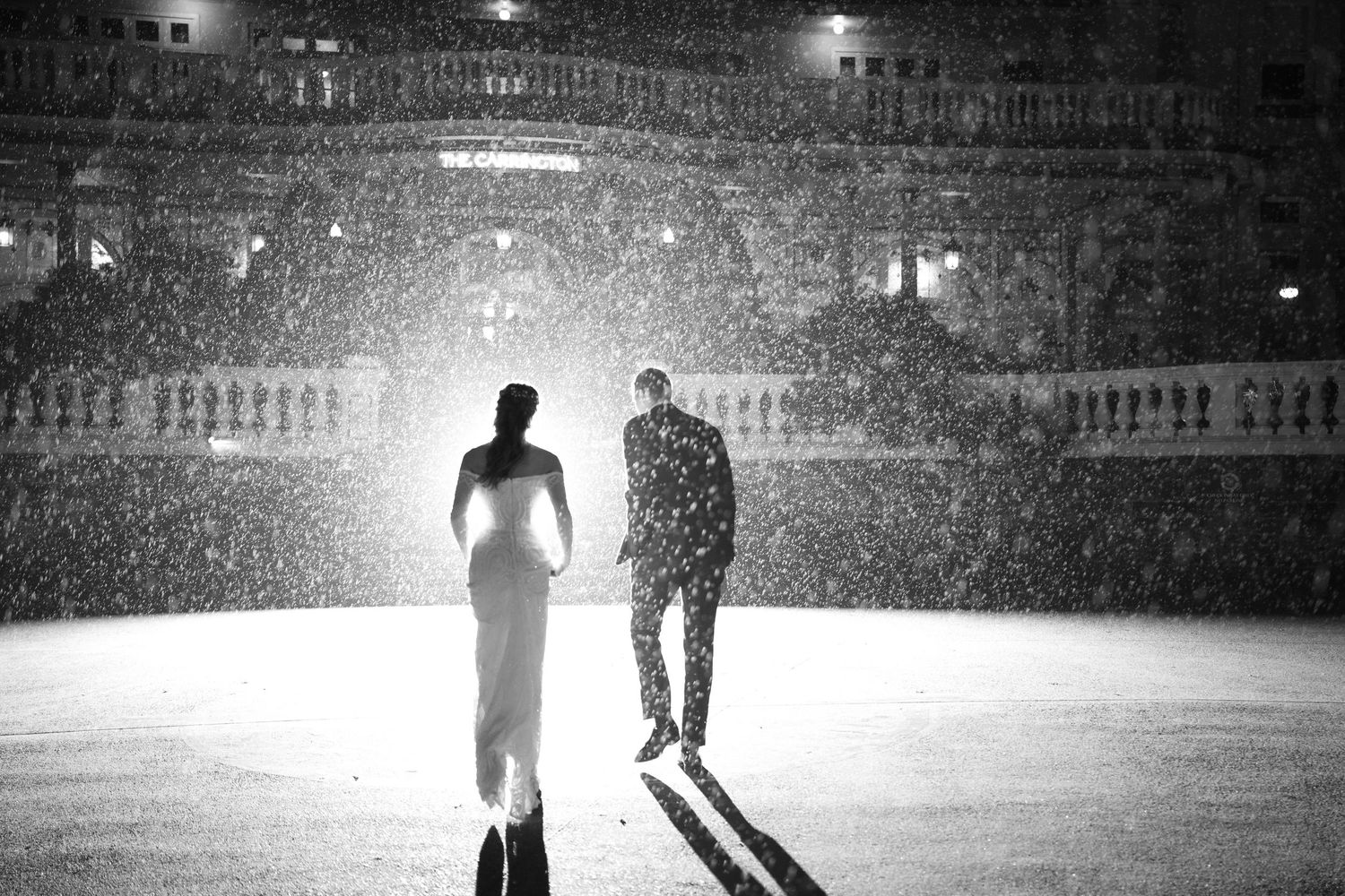 Black and white photo of a couple standing in the snow at night, with a grand building illuminated behind them.