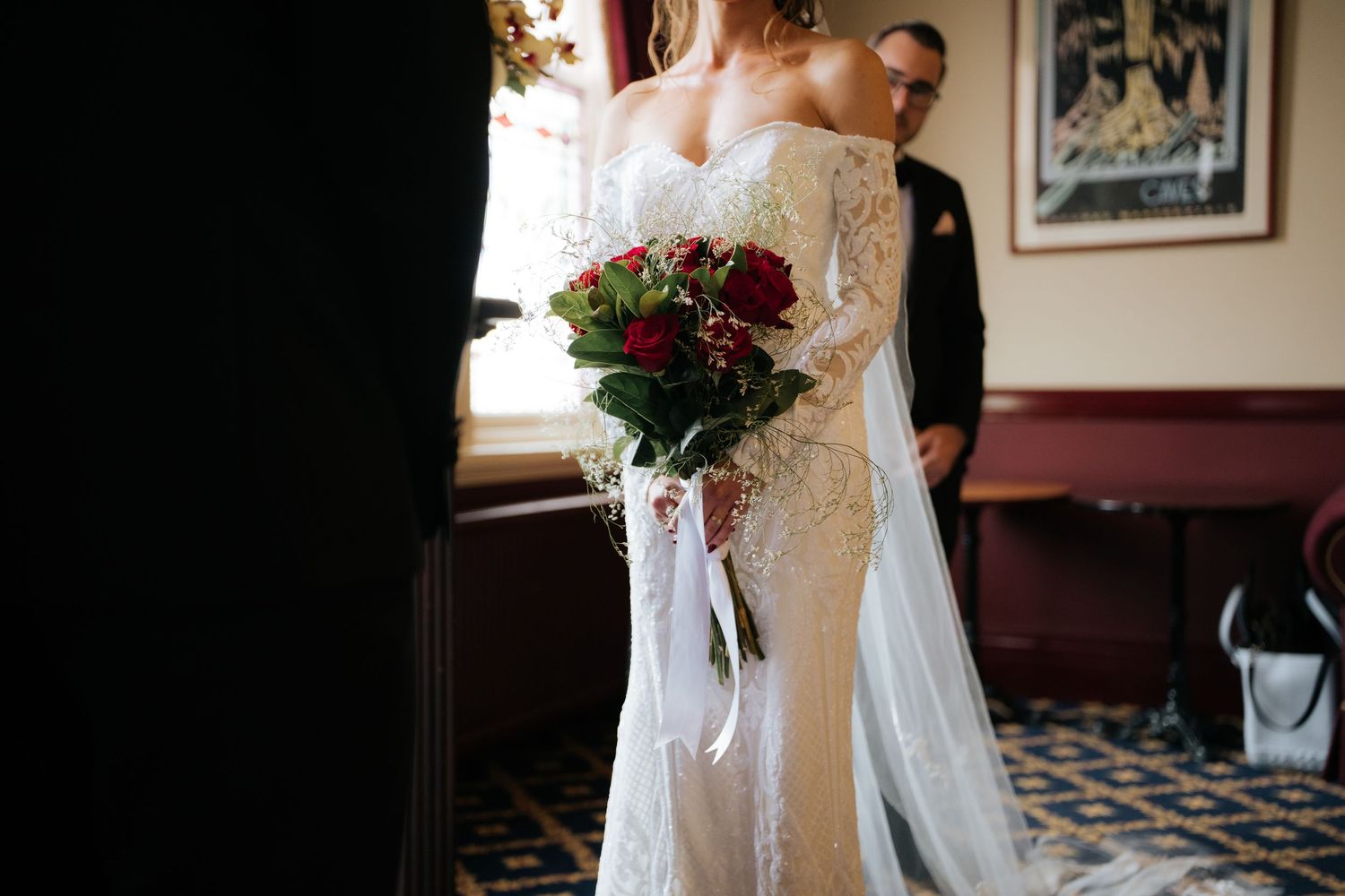 Bride in lace wedding gown holding red rose bouquet with white ribbons in elegant venue.