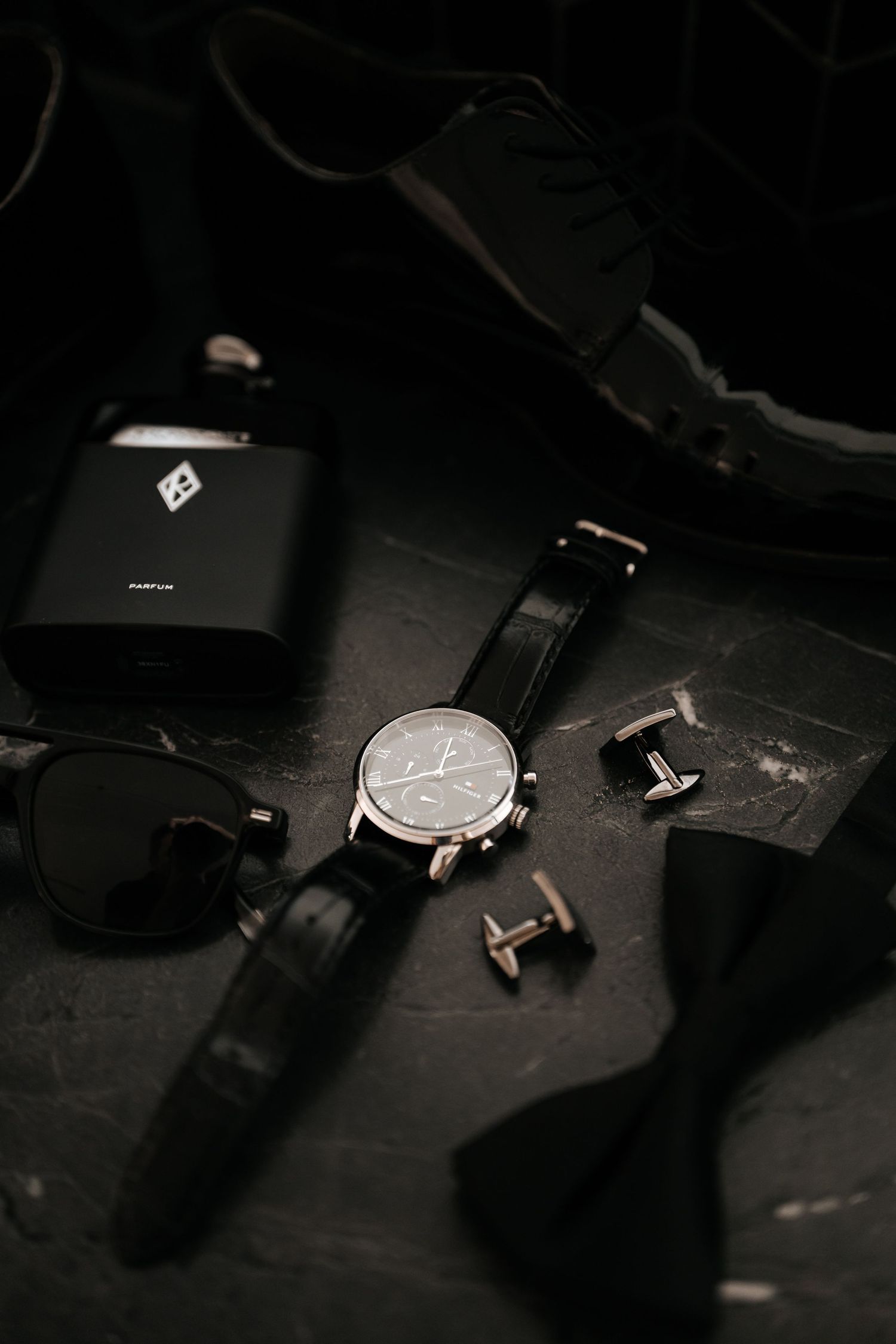 Groom wedding accessories flatlay featuring a classic watch, cufflinks, sunglasses, flask, and dress shoes on dark surface.
