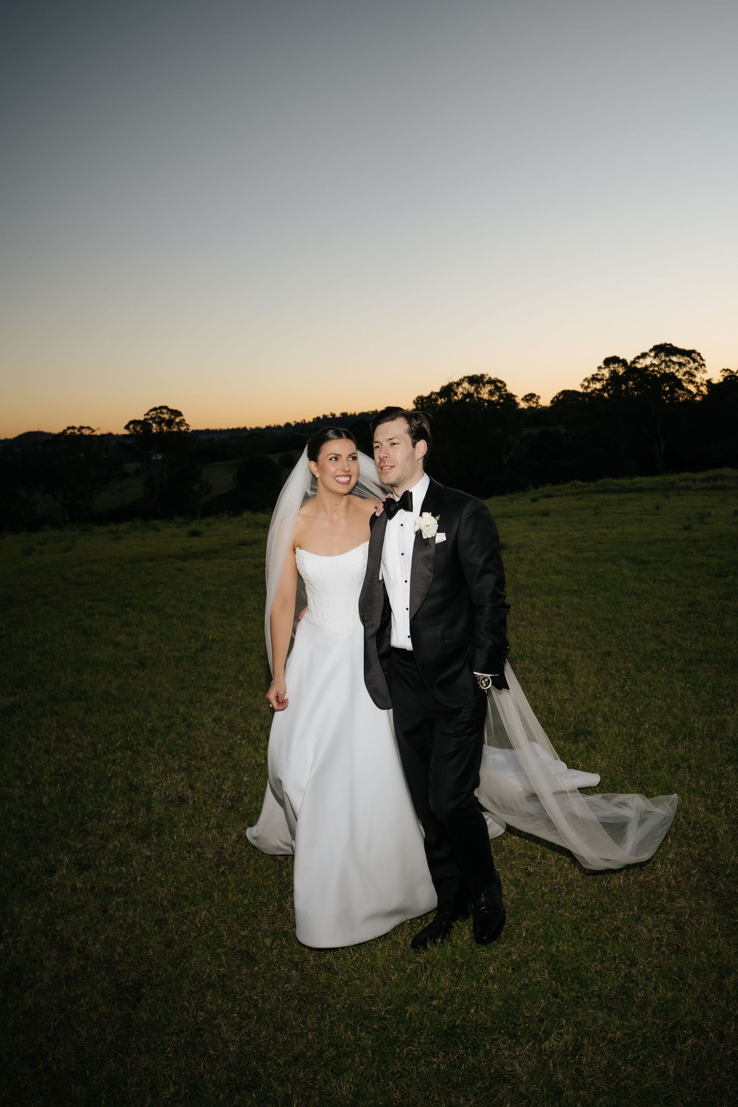 Smiling bride and groom in tuxedo pose together on green lawn at dusk during wedding celebration.