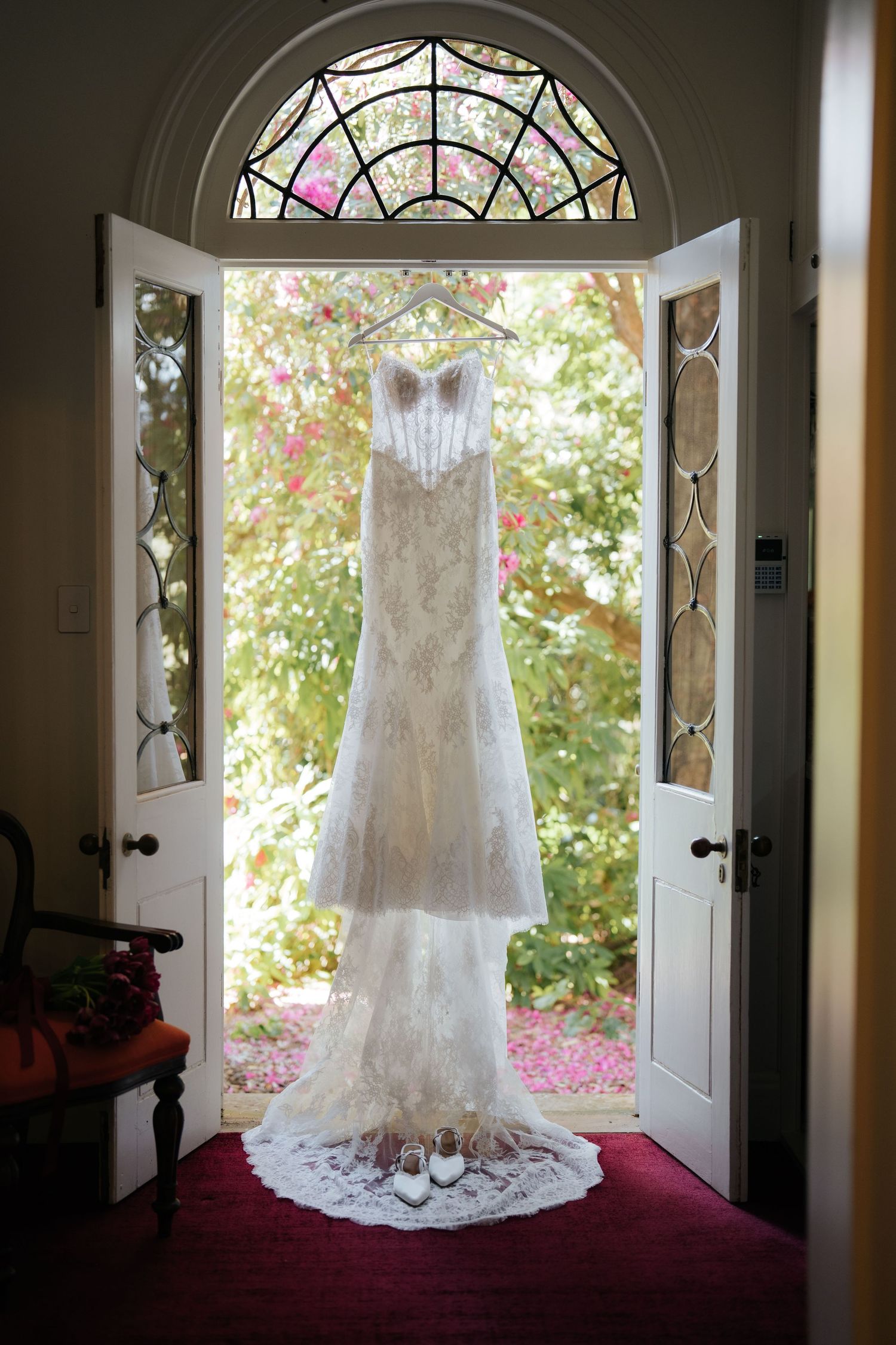 Elegant lace mermaid wedding dress hanging in a doorway with stained glass windows and bridal shoes below.