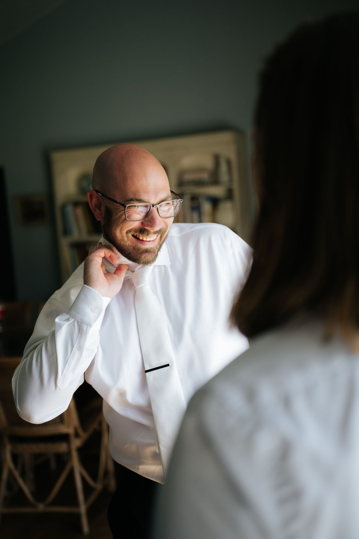 Smiling bald groom in white dress shirt adjusting bow tie while getting ready for wedding day.