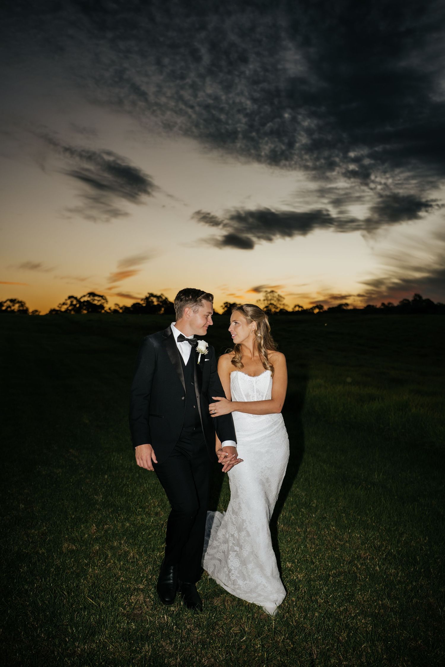 Bride and groom sharing a romantic moment at sunset on a lush green field under a dramatic cloudy sky.