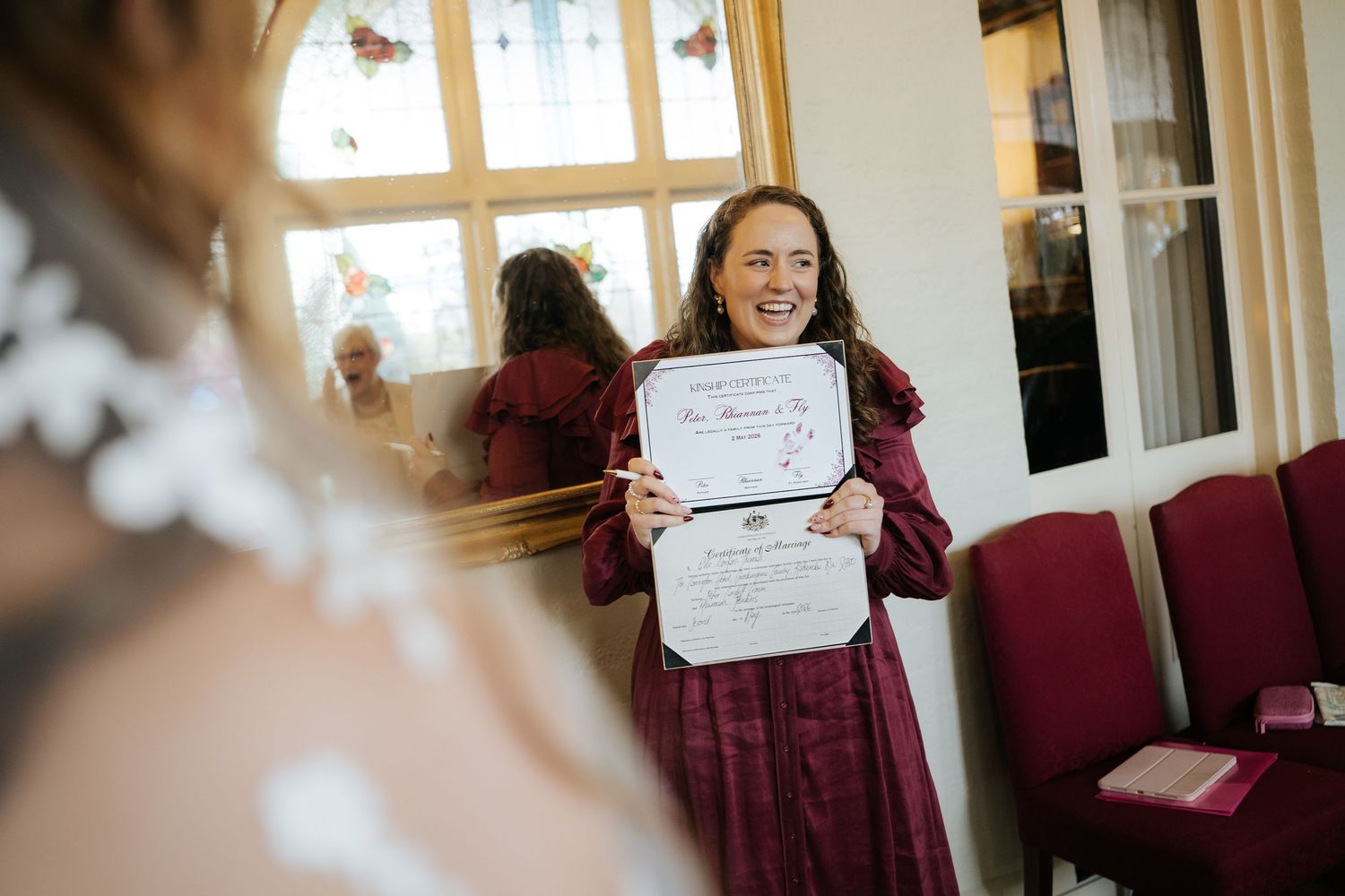Woman in burgundy dress proudly displays a wedding certificate, smiling at the bride during a wedding ceremony.