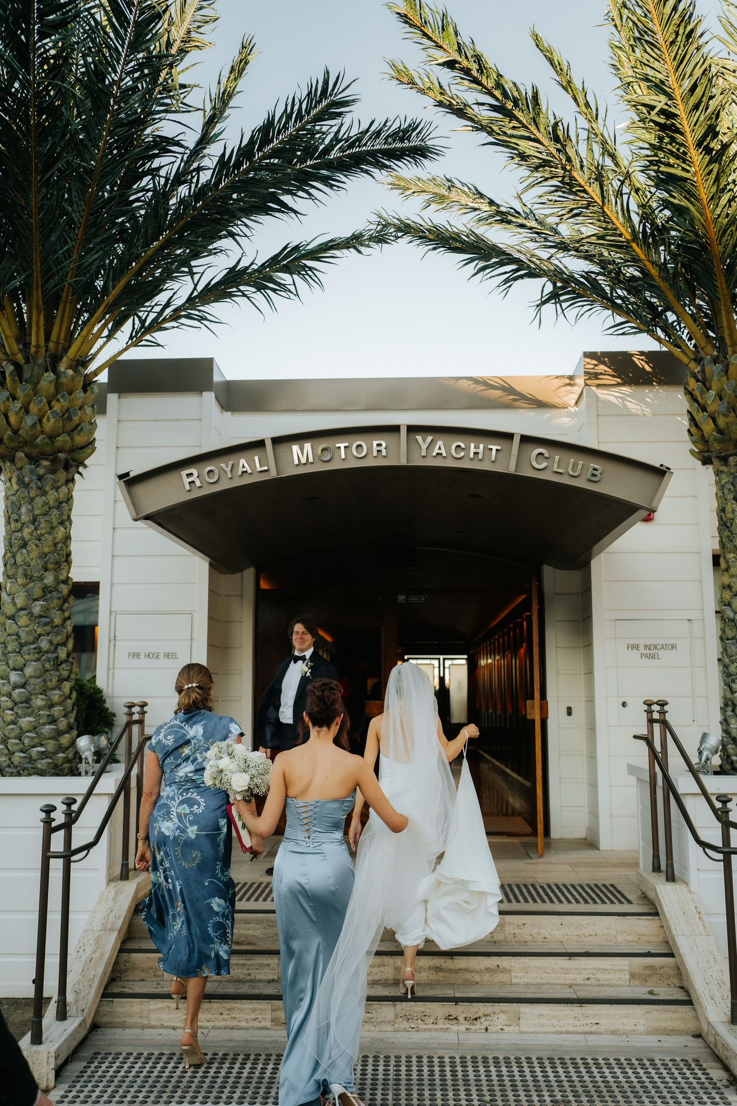 Bride and wedding party ascending steps to Royal Motor Yacht Club flanked by palm trees.