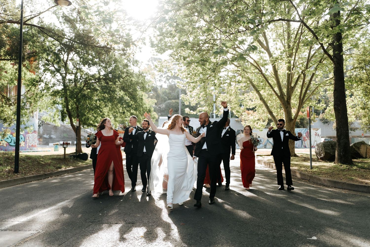 Wedding party celebrating outdoors, bride and groom leading group down tree-lined street, bridesmaids in red dresses.