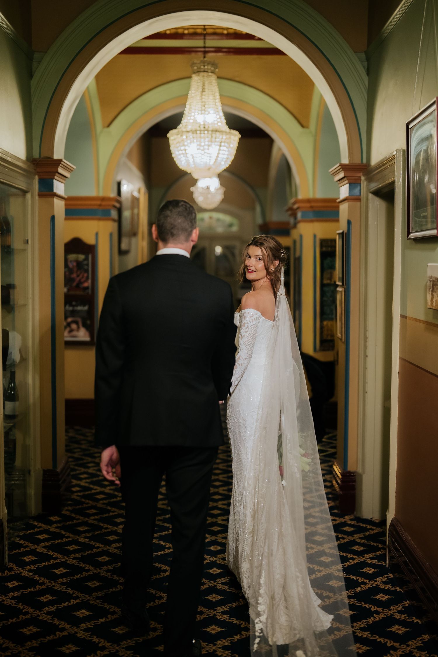 Bride in white lace gown with veil looks back at groom in black suit in elegant hallway with chandelier.