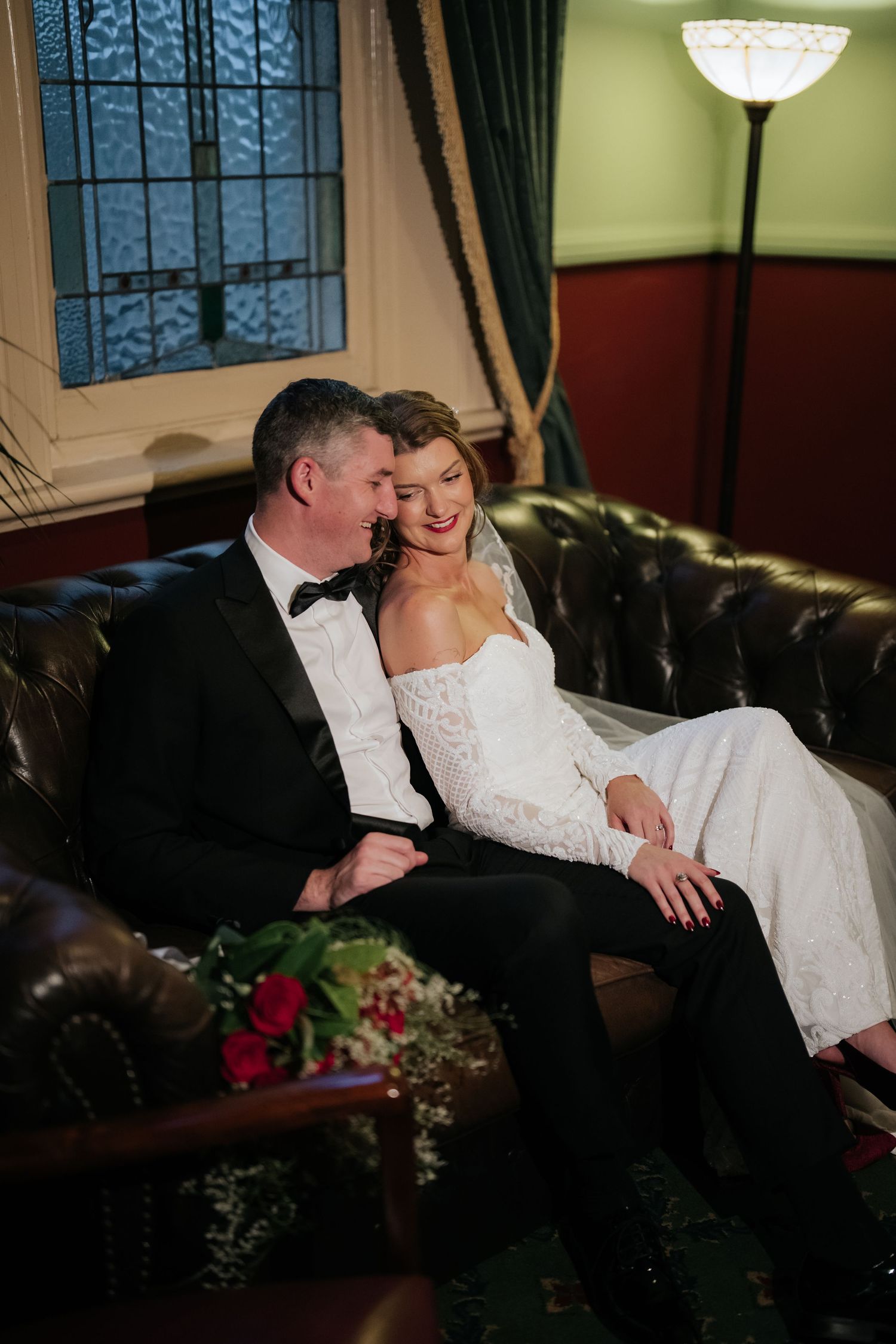 A bride and groom share a romantic moment seated on a leather sofa, dressed in wedding attire with a bouquet nearby.