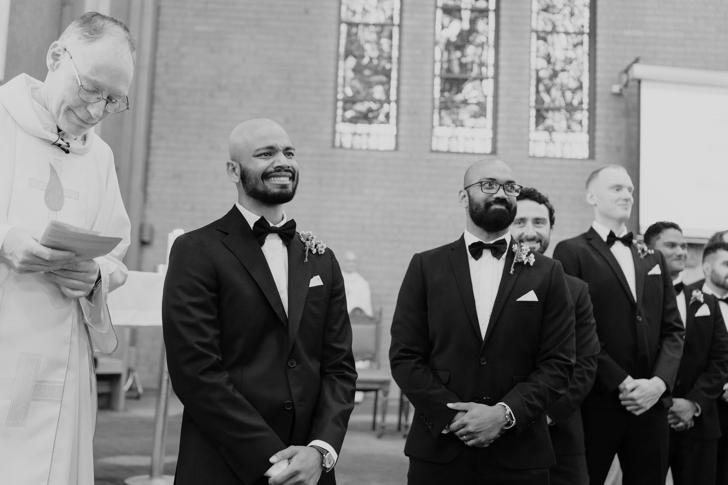 Groom and groomsmen smiling at the altar during a church wedding ceremony in black and white photo.