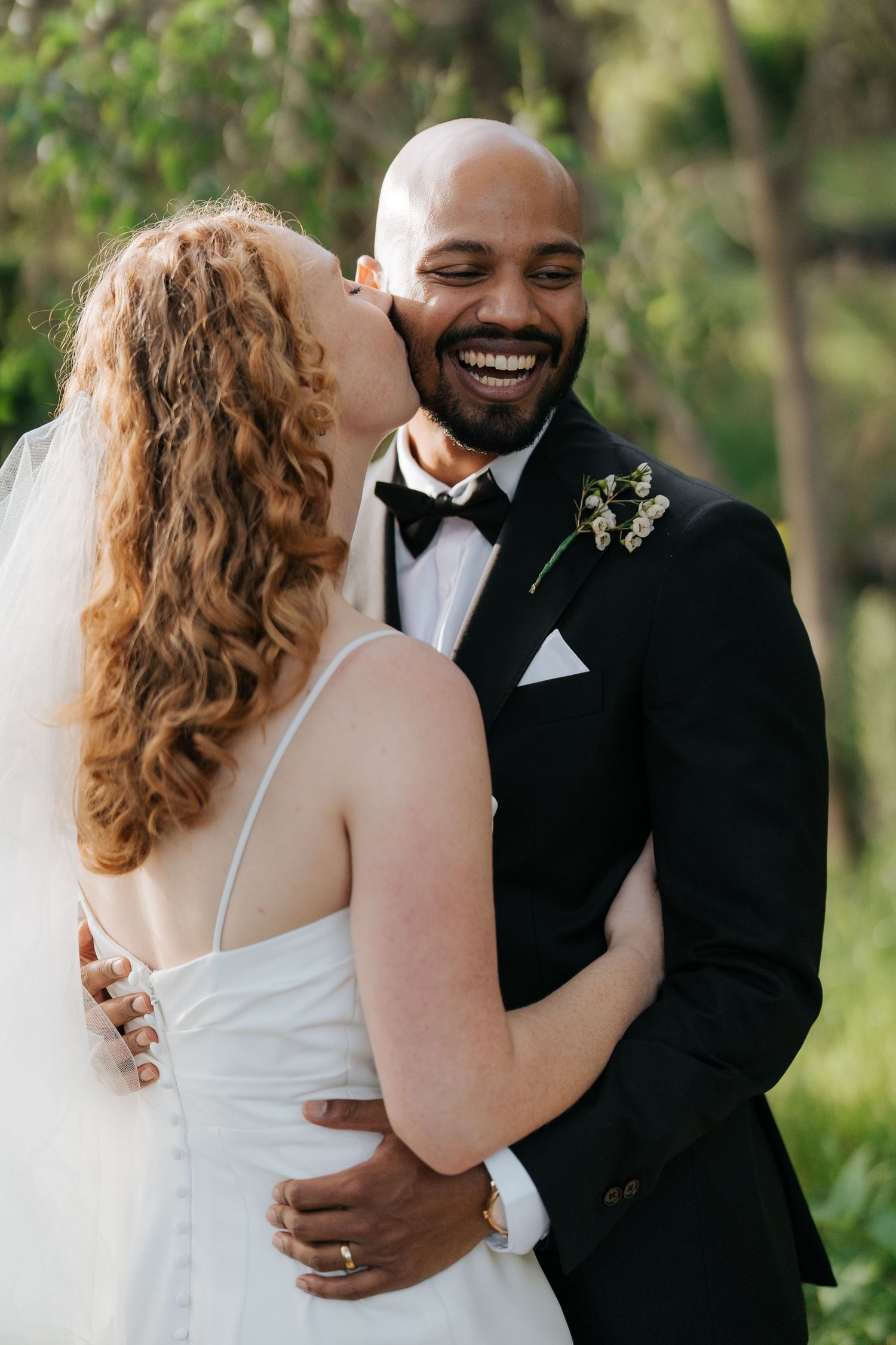 A bride in a white gown kisses a smiling groom in a black tuxedo outdoors on their wedding day.