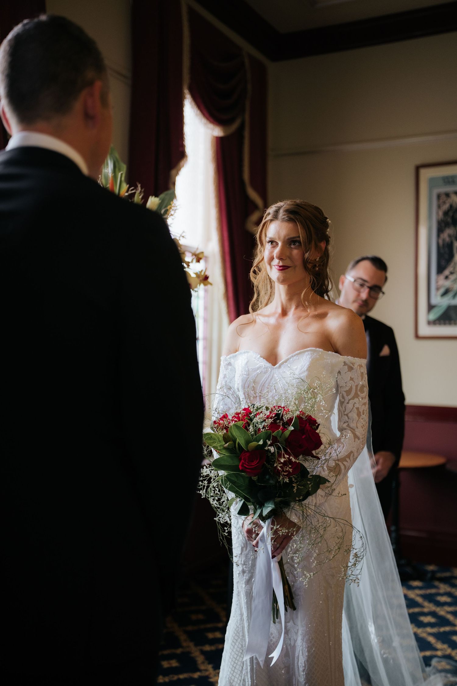 Bride in lace off-shoulder gown holds red rose bouquet during indoor wedding ceremony.