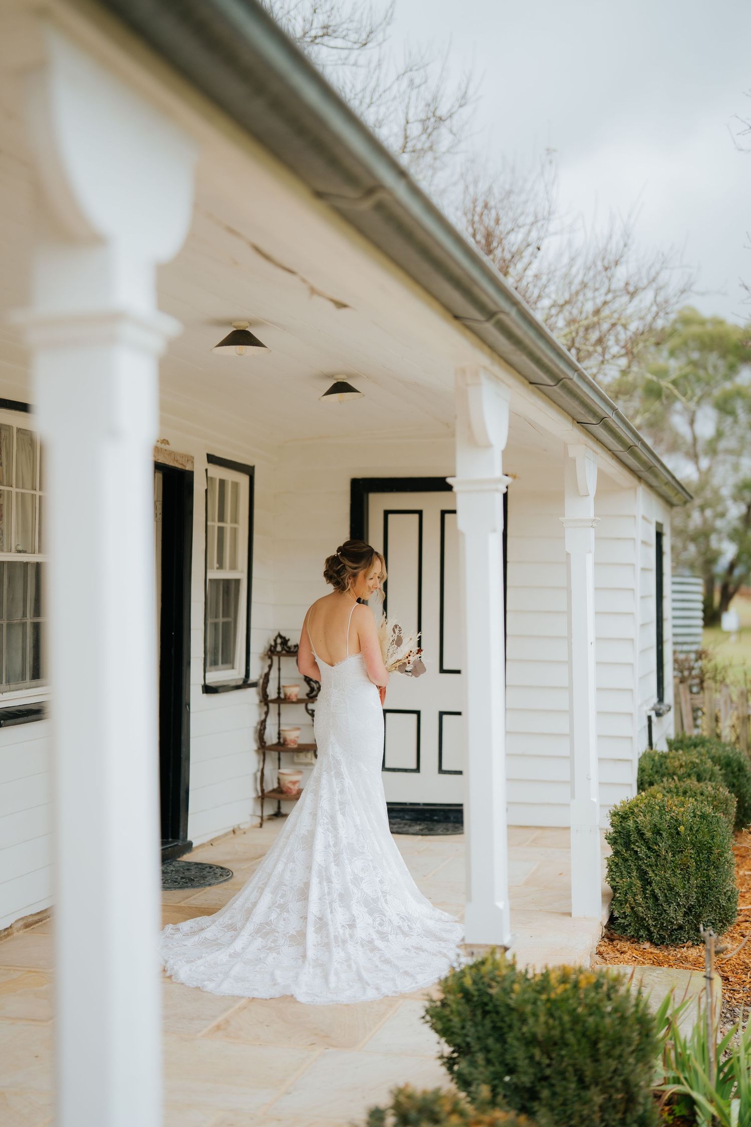 Bride in lace wedding gown with train stands on white colonial porch holding champagne glass.