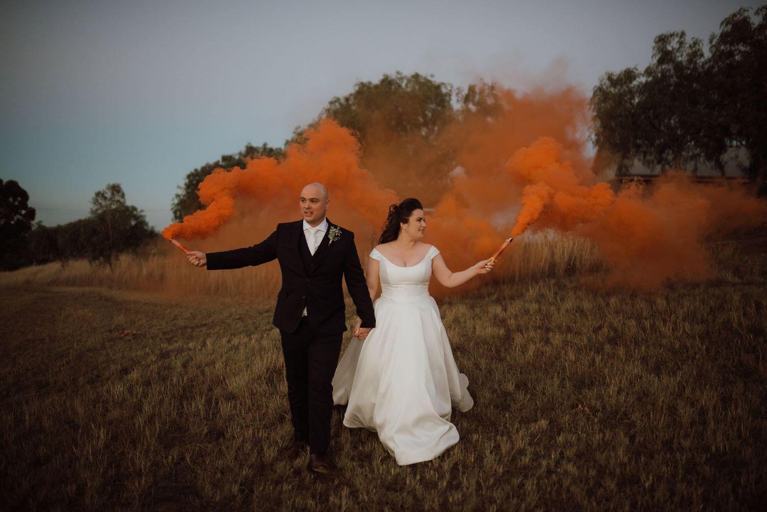 Bride and groom holding orange smoke bombs in a field, creating a dramatic and colorful wedding photo.