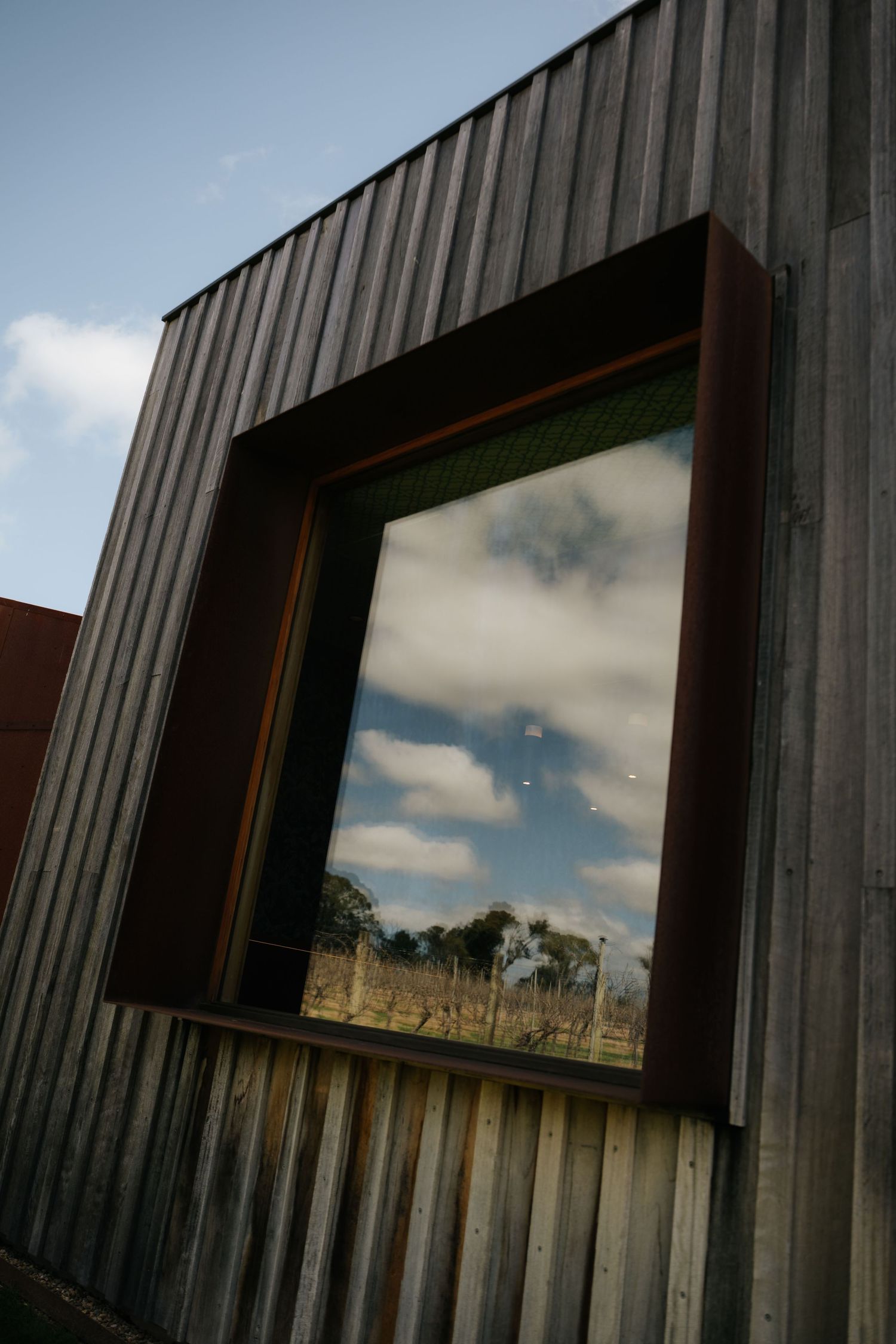 Corrugated metal building exterior with a large window reflecting clouds and trees against a blue sky.