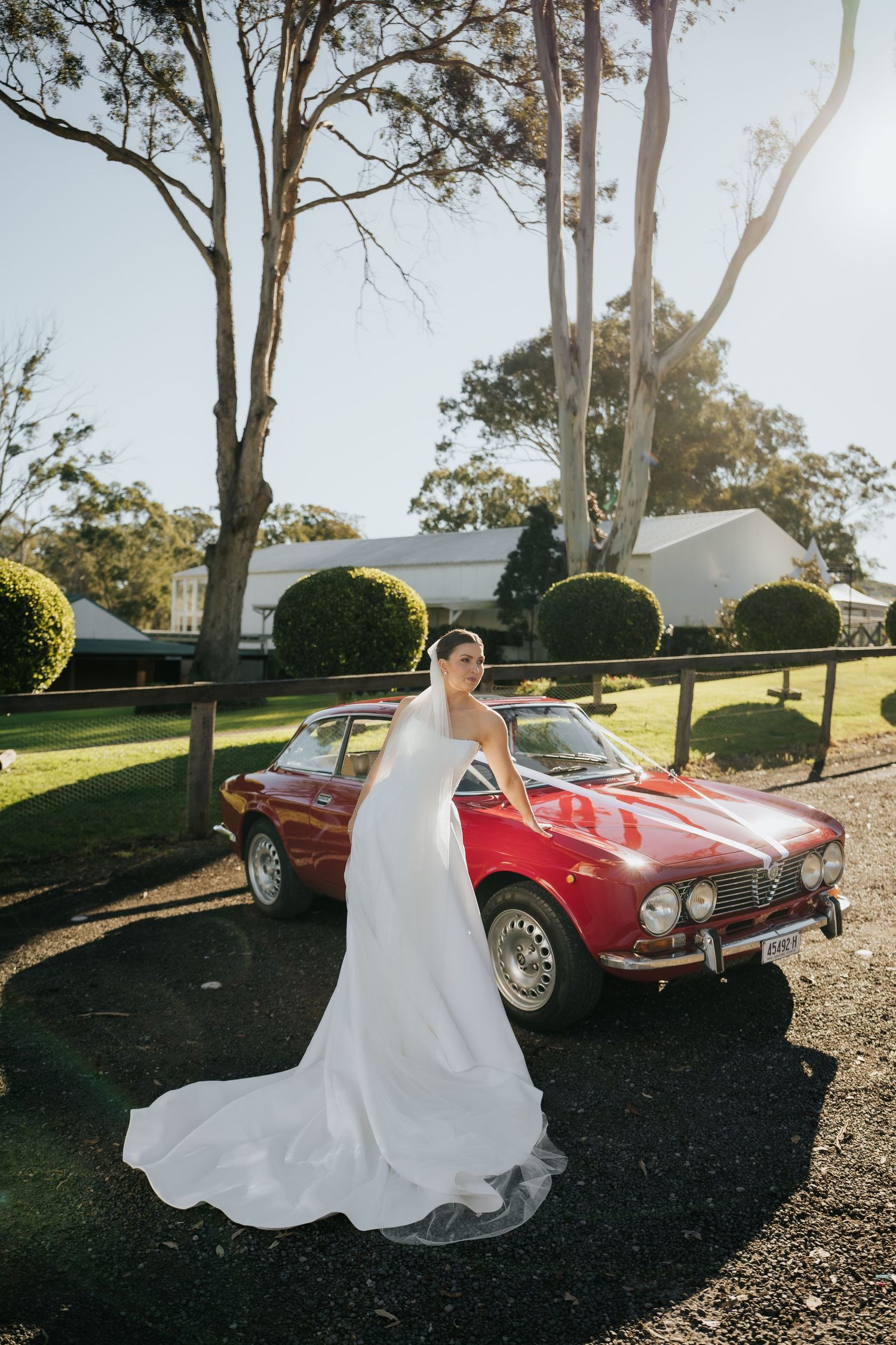 Elegant bride in strapless white gown with long train posing beside a classic red vintage car outdoors.