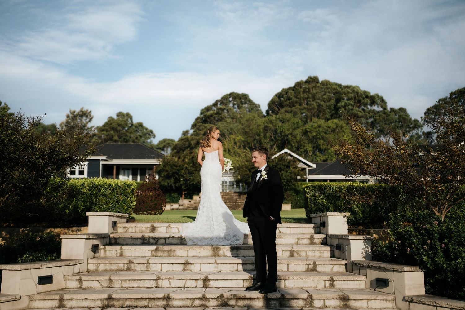 Bride and groom pose on stone steps surrounded by lush greenery at an elegant outdoor wedding venue.