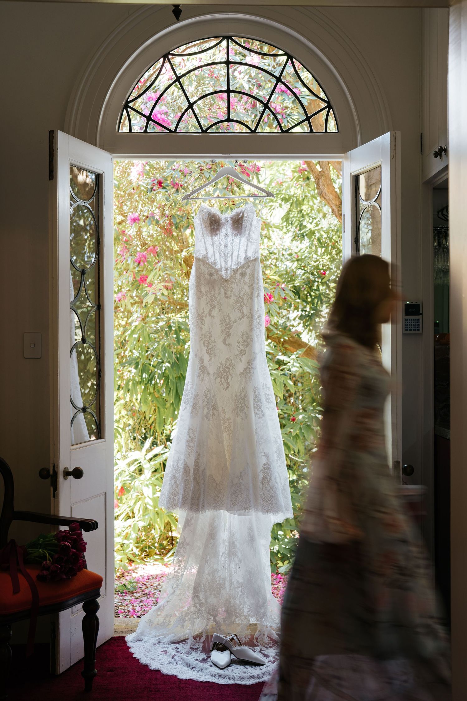 Elegant lace wedding dress hanging in a sunlit doorway with stained glass arch, bridal shoes below, and blurred figure nearby.