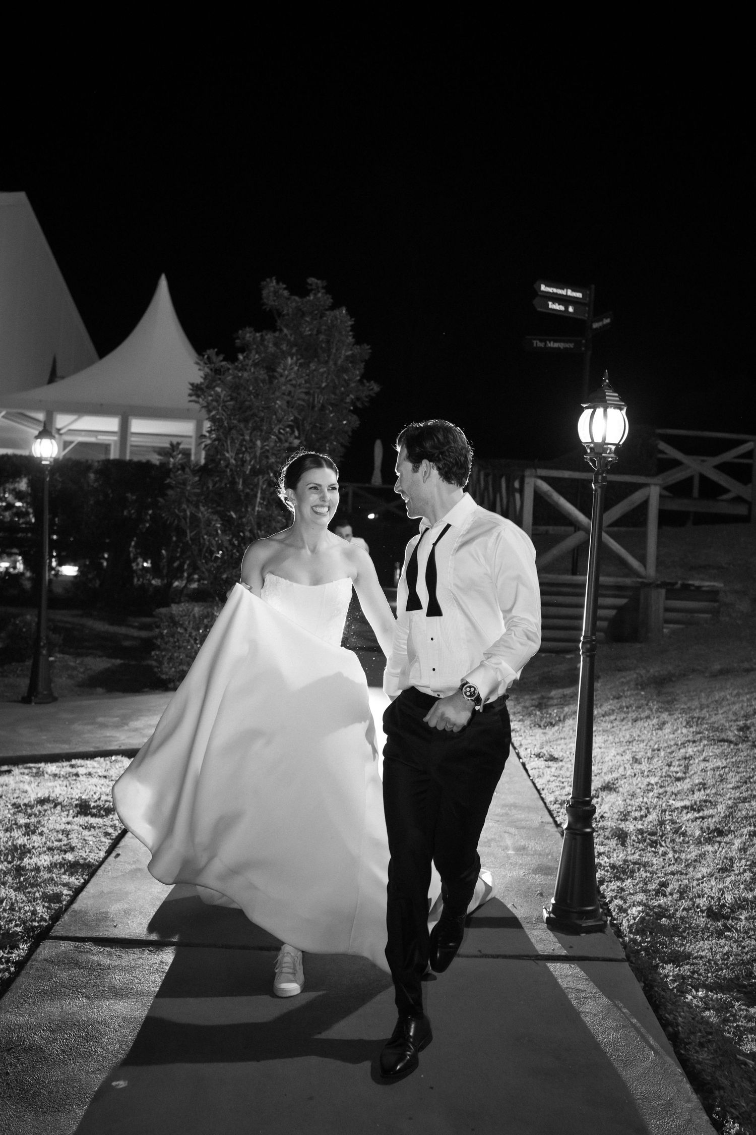 Bride and groom walk together at night outdoors, smiling, in black and white wedding photo.
