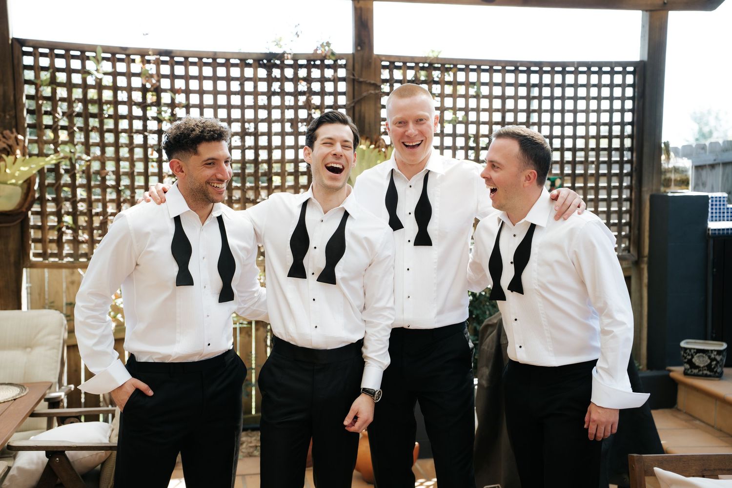 Four groomsmen in white shirts and black bow ties laughing together before a wedding ceremony.