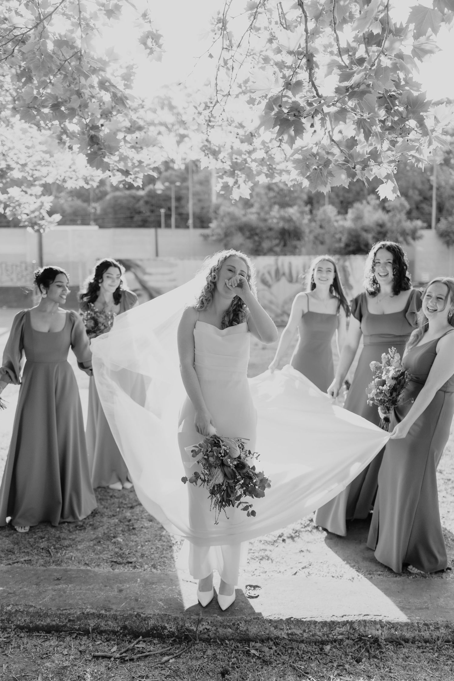 Black and white photo of a bride holding a bouquet with her veil flowing, surrounded by smiling bridesmaids outdoors.
