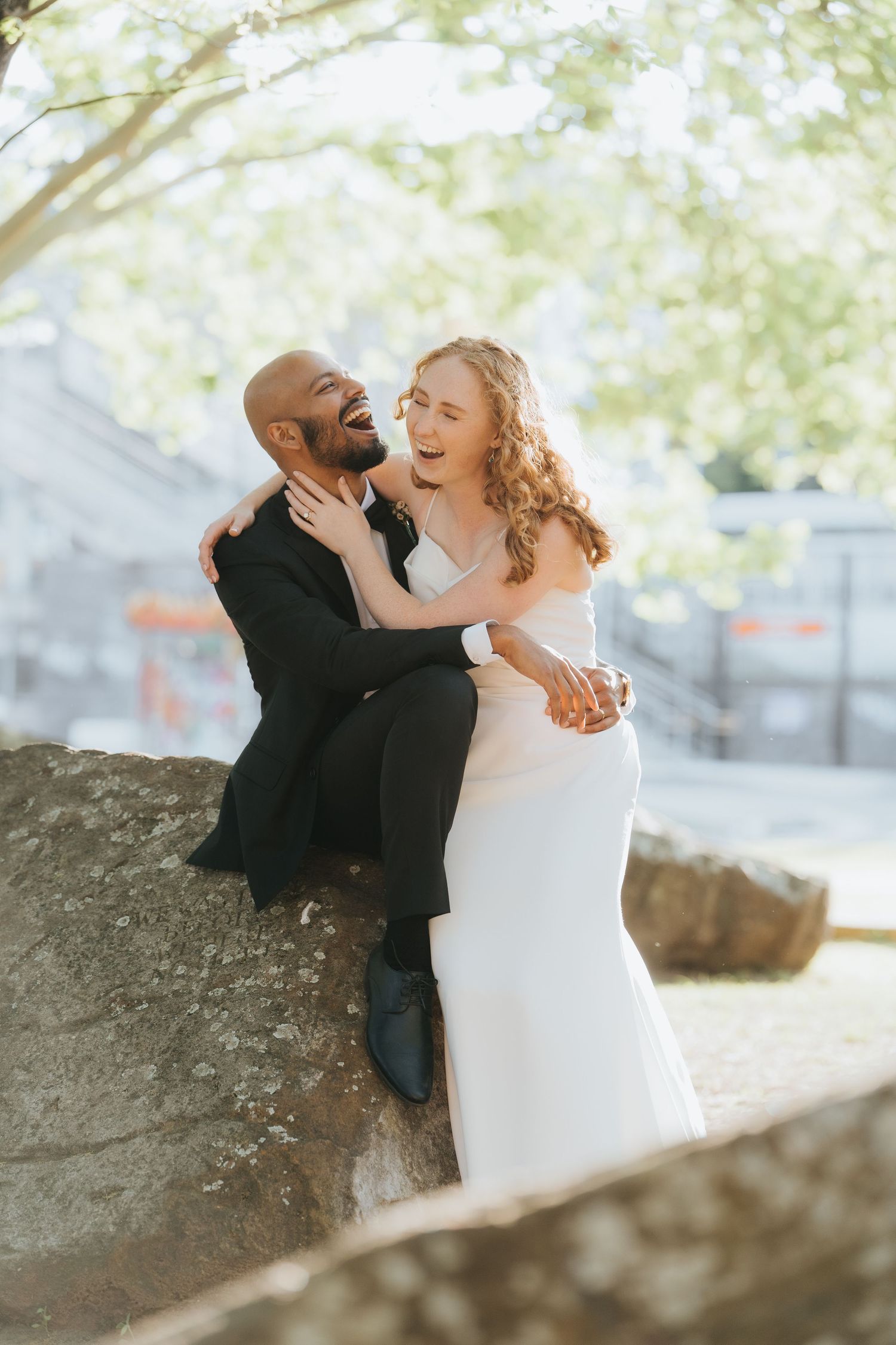 A joyful interracial couple in wedding attire laugh together outdoors, bride in white gown embracing suited groom on a rock.