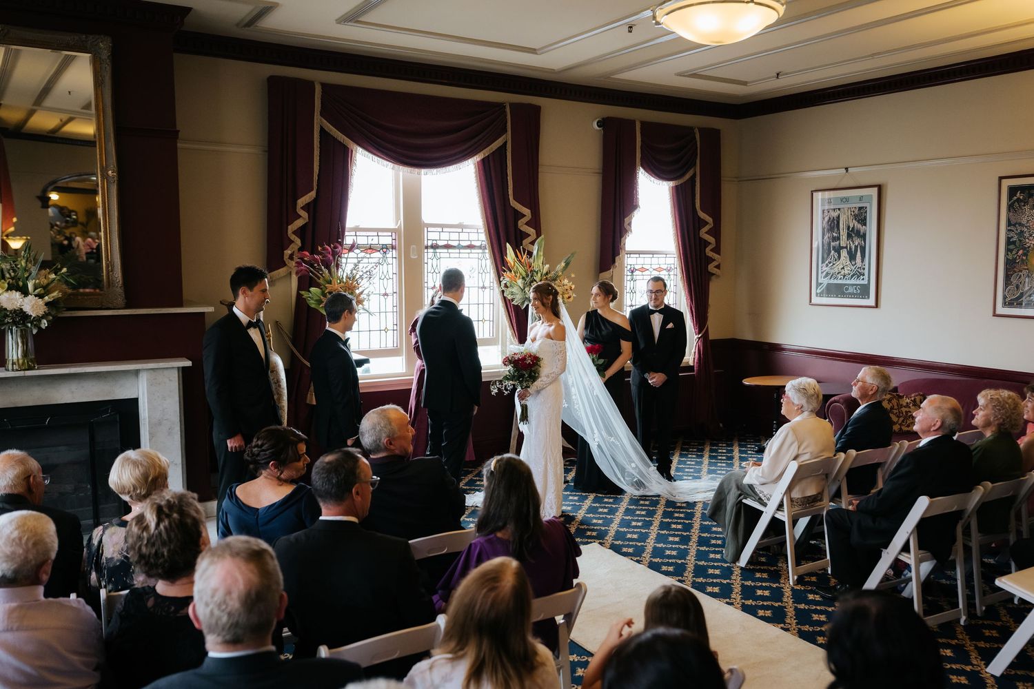 Elegant indoor wedding ceremony with bride and groom at altar, guests seated in white chairs, ornate drapes and floral arrangements.