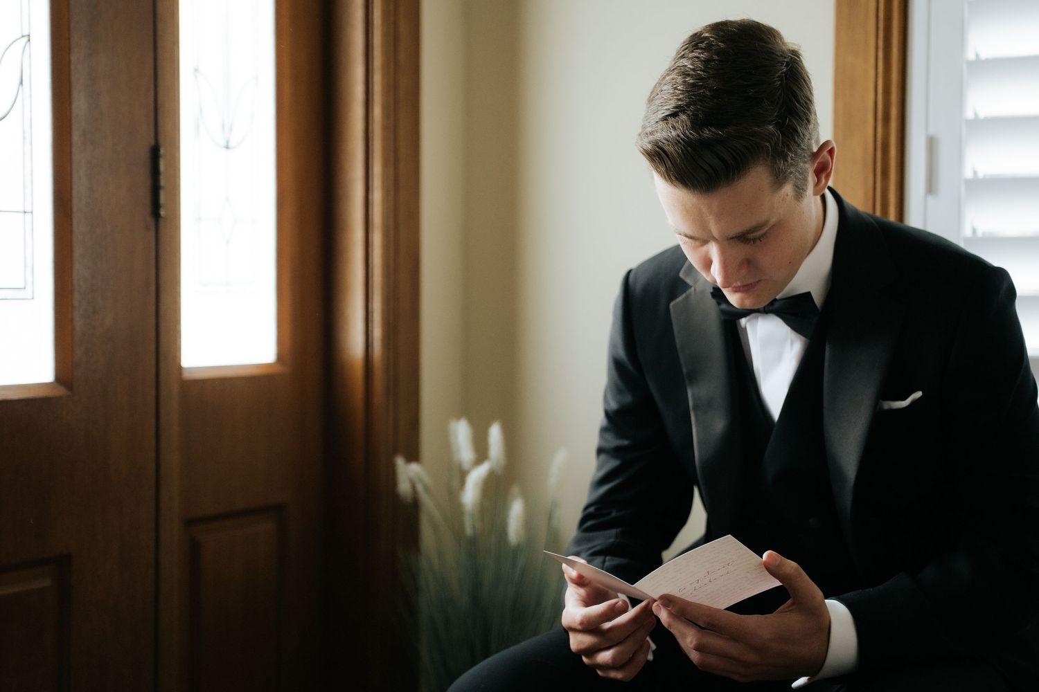 A groom in a black tuxedo and bow tie reads a heartfelt note while seated near a window before his wedding ceremony.