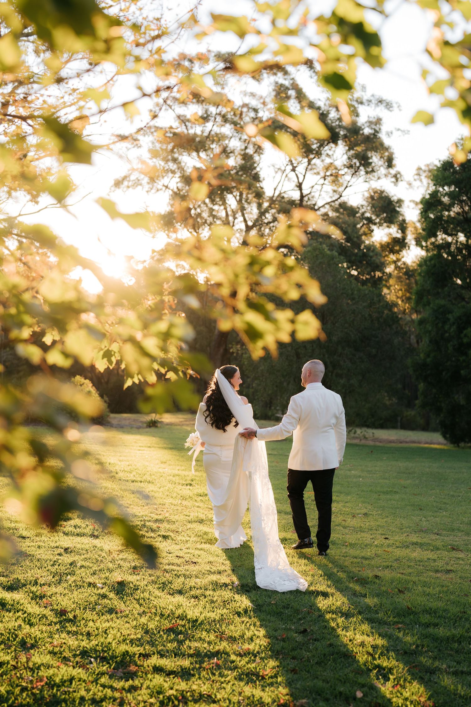 Bride and groom holding hands walking through sunlit green garden at golden hour wedding.