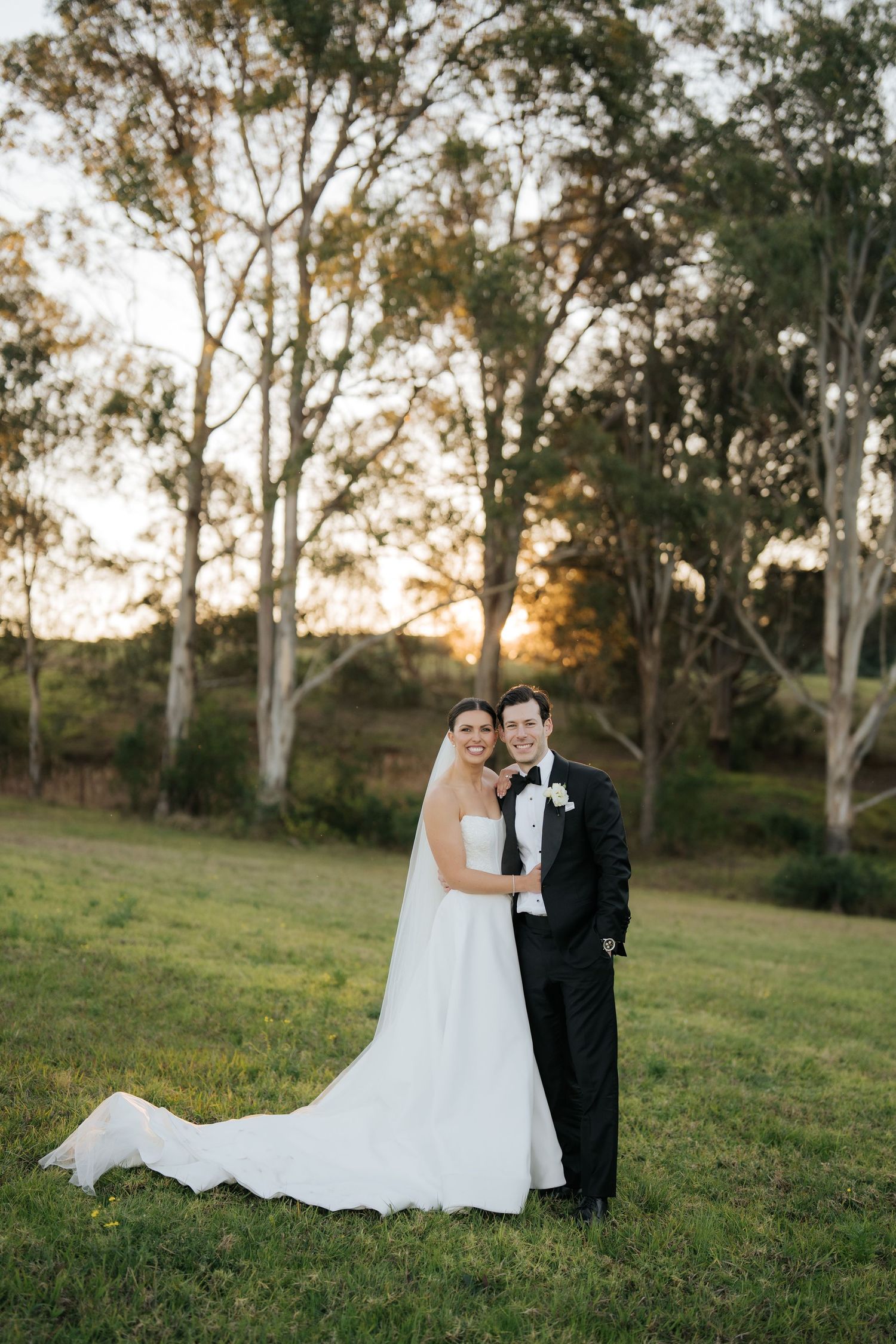 Bride and groom embracing in green field at sunset, bride in white gown, groom in black tuxedo.