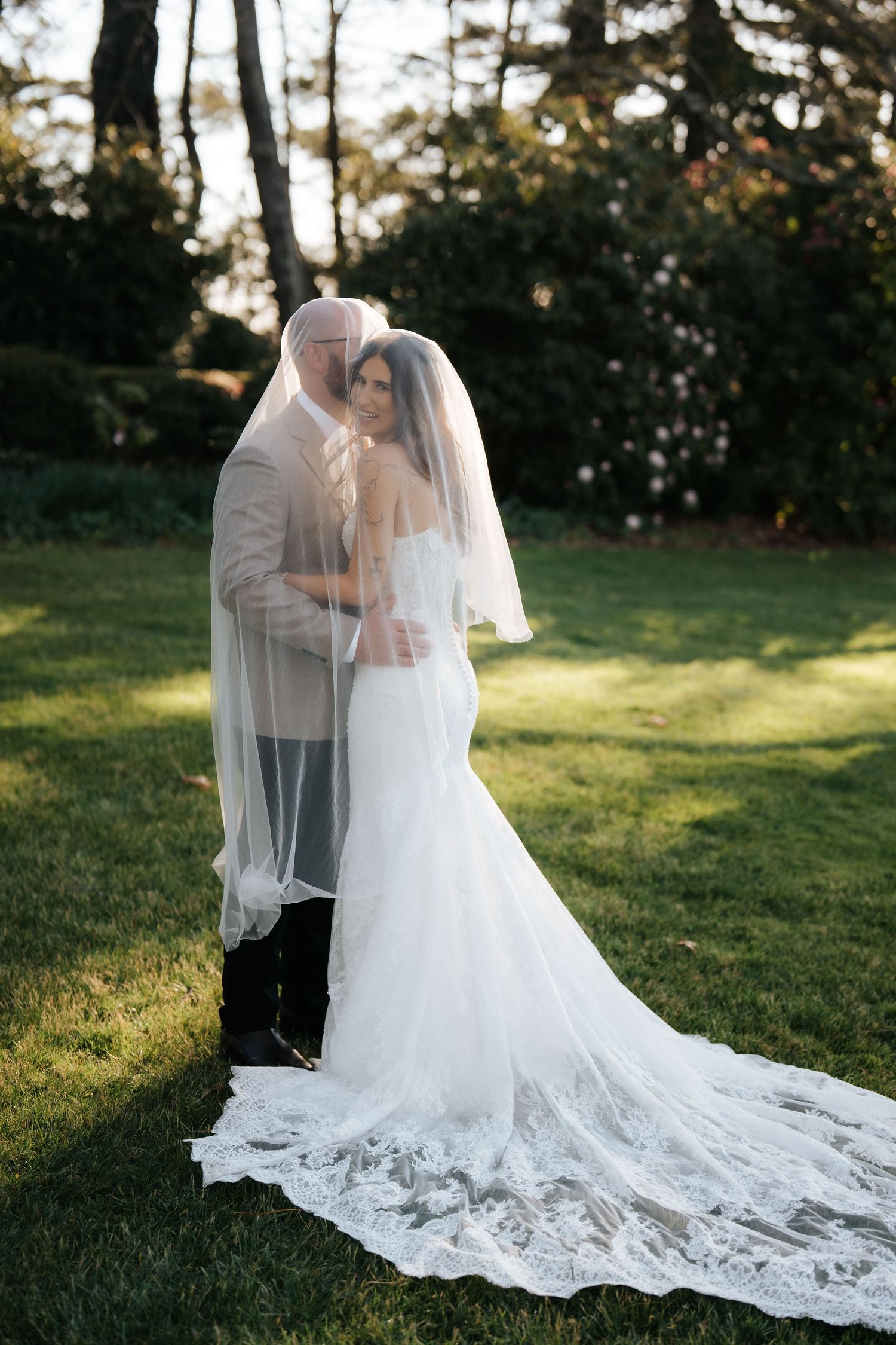 Bride and groom share a romantic moment under a flowing lace veil in a lush green garden during golden hour.