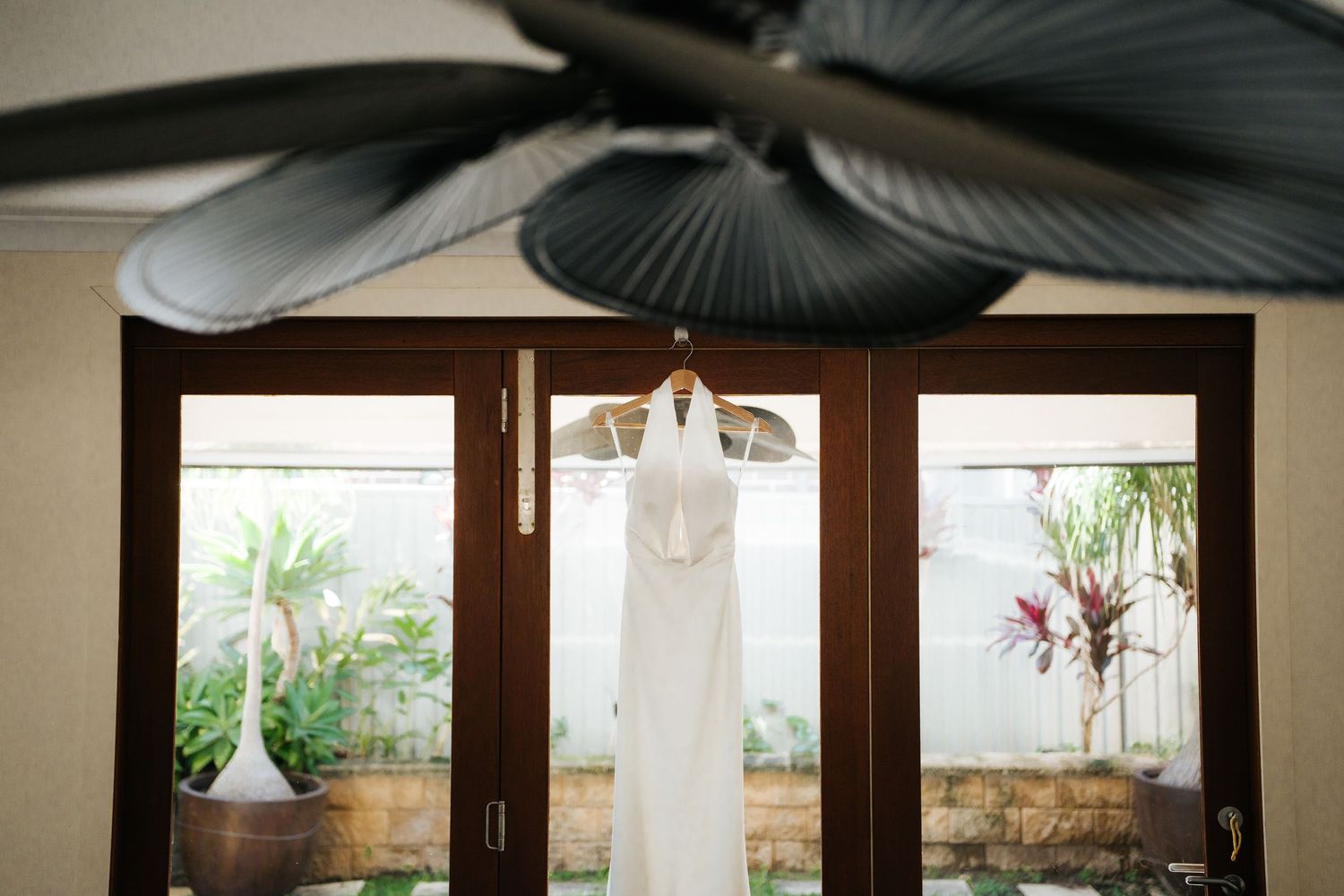 White wedding dress hanging on wooden French doors with tropical garden view and ceiling fan above.