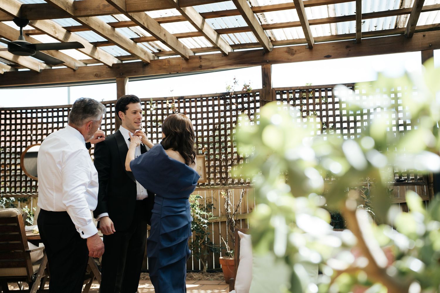 Man in suit gets tie adjusted by woman on pergola patio while another man in white shirt watches.