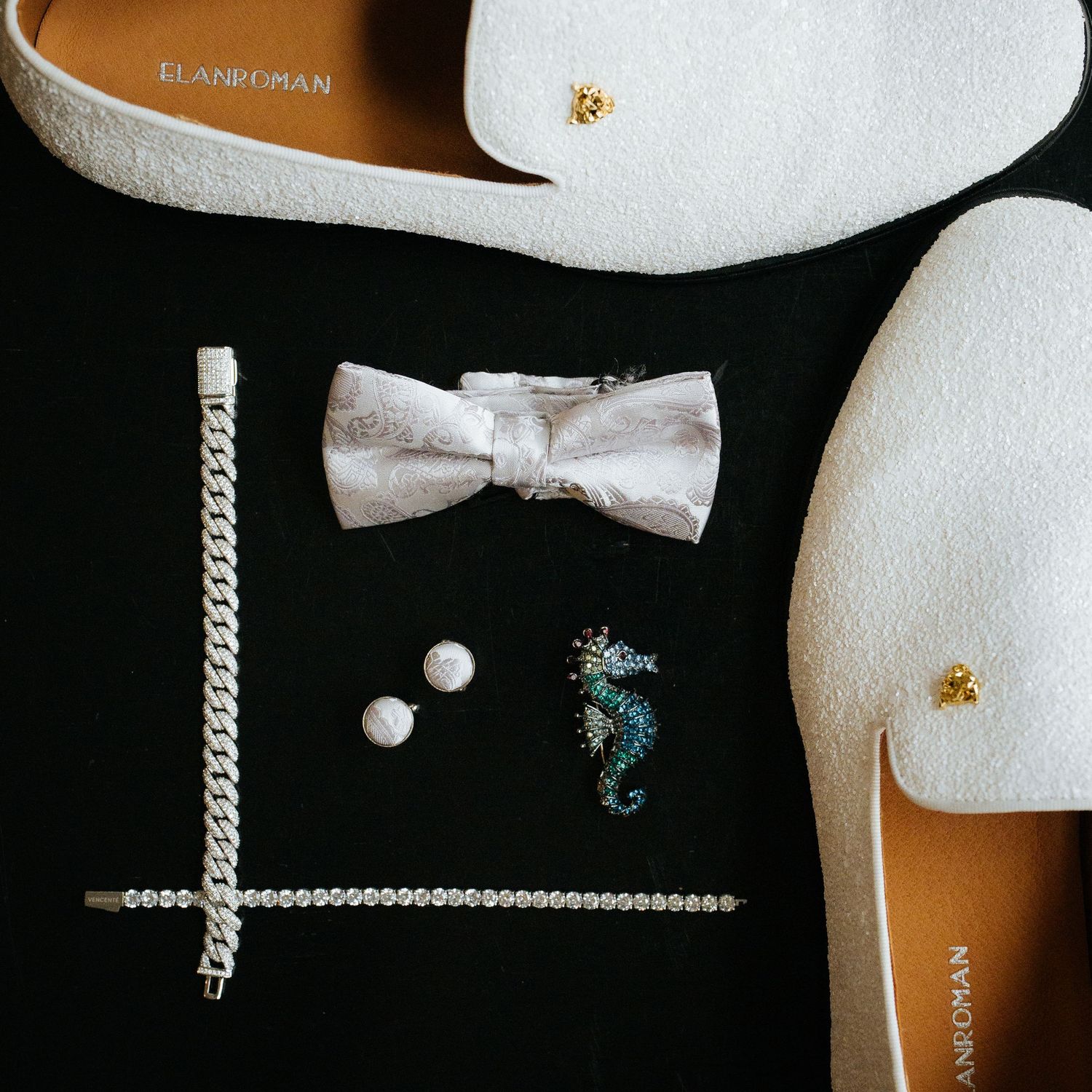 Flat lay of white wedding accessories including bow tie, bracelet, earrings and loafers.