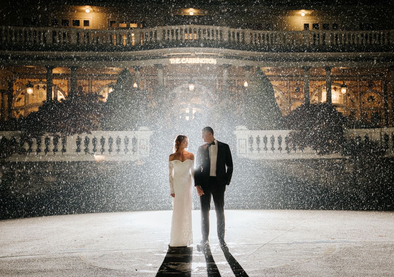 Couple in wedding attire standing in snow at night outside an illuminated classical building.